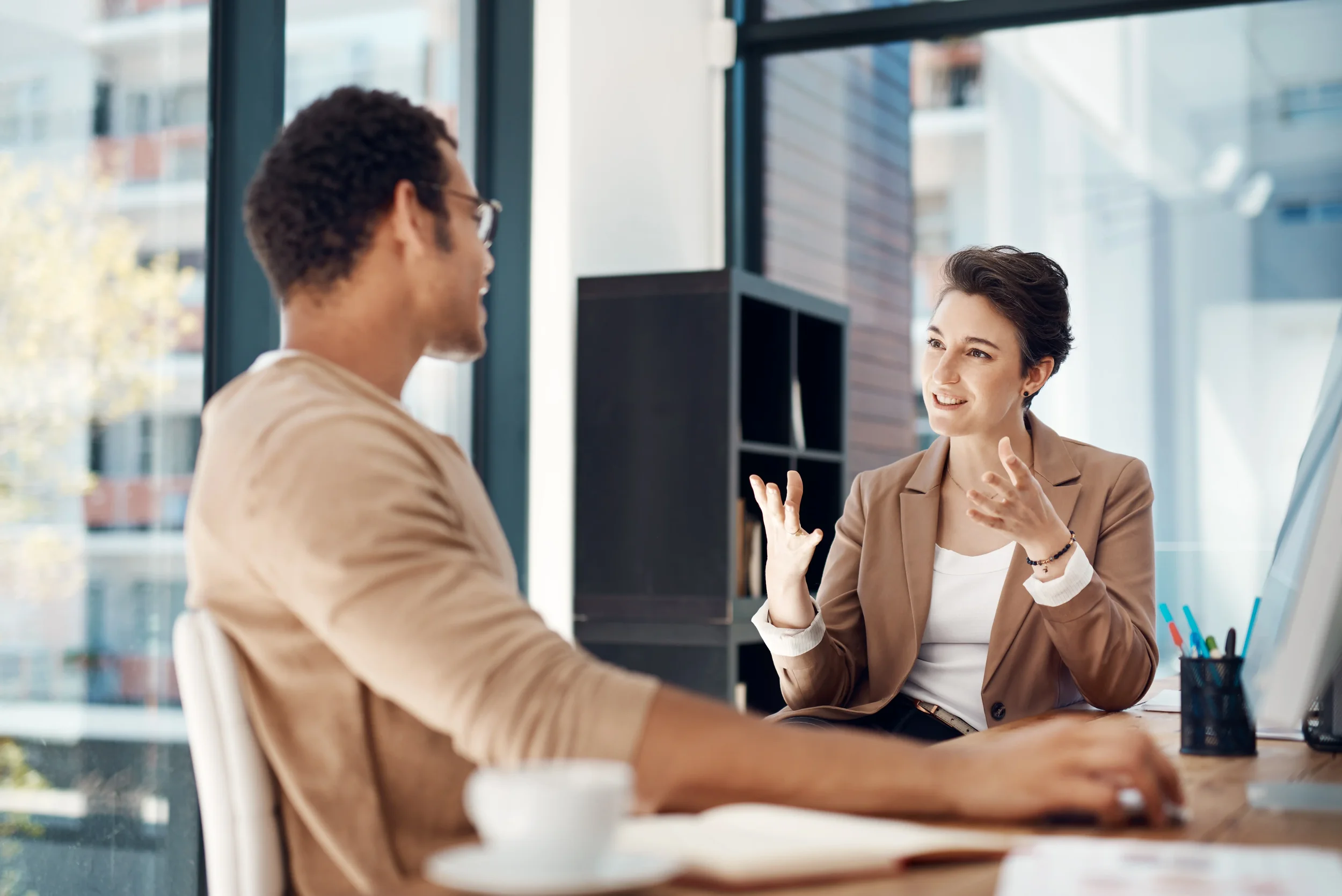 Two professional services colleagues in conversation at a modern office desk, discussing strategy over coffee