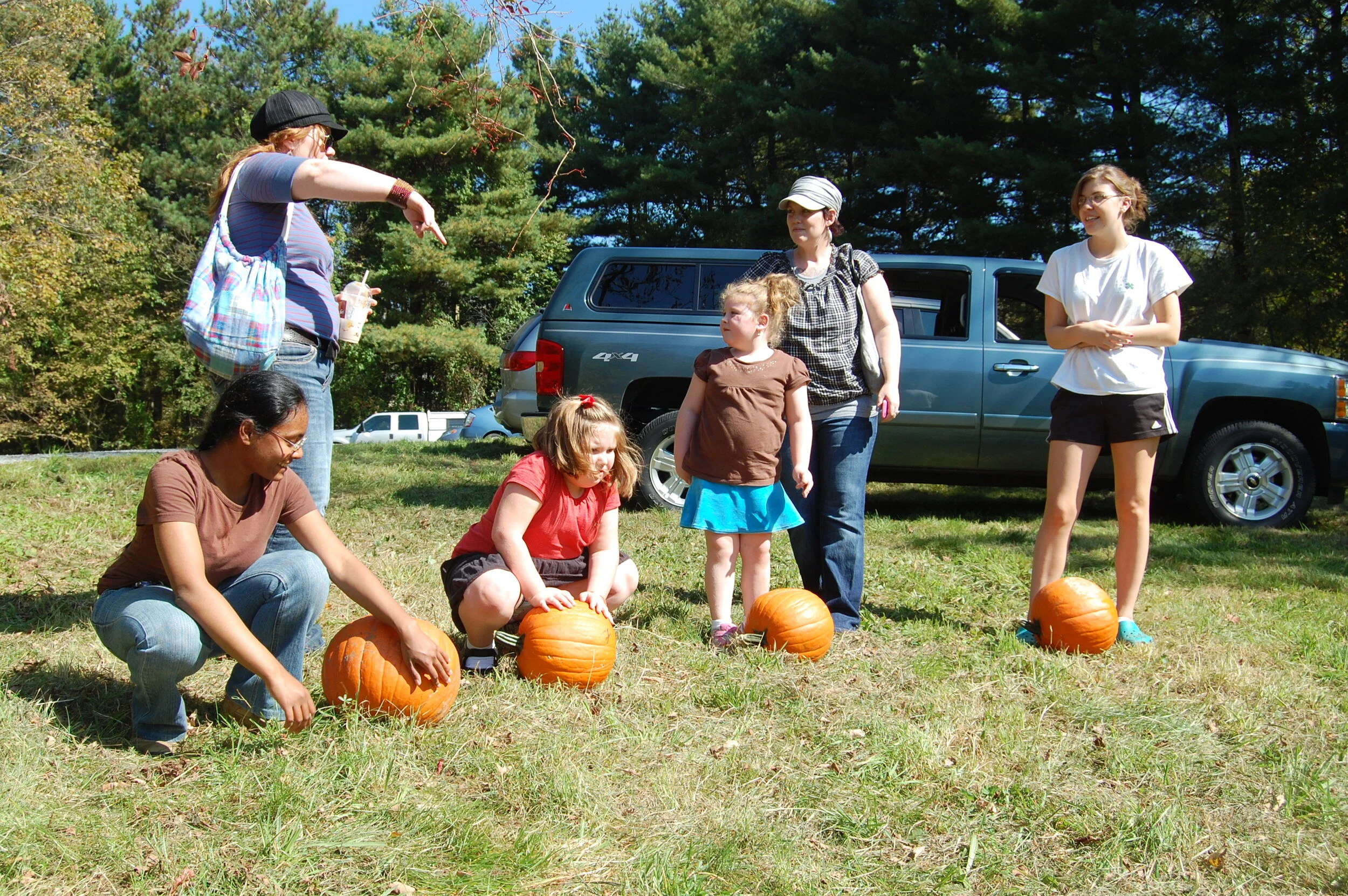 Group of people outdoors with pumpkins on grass, SUV in background.