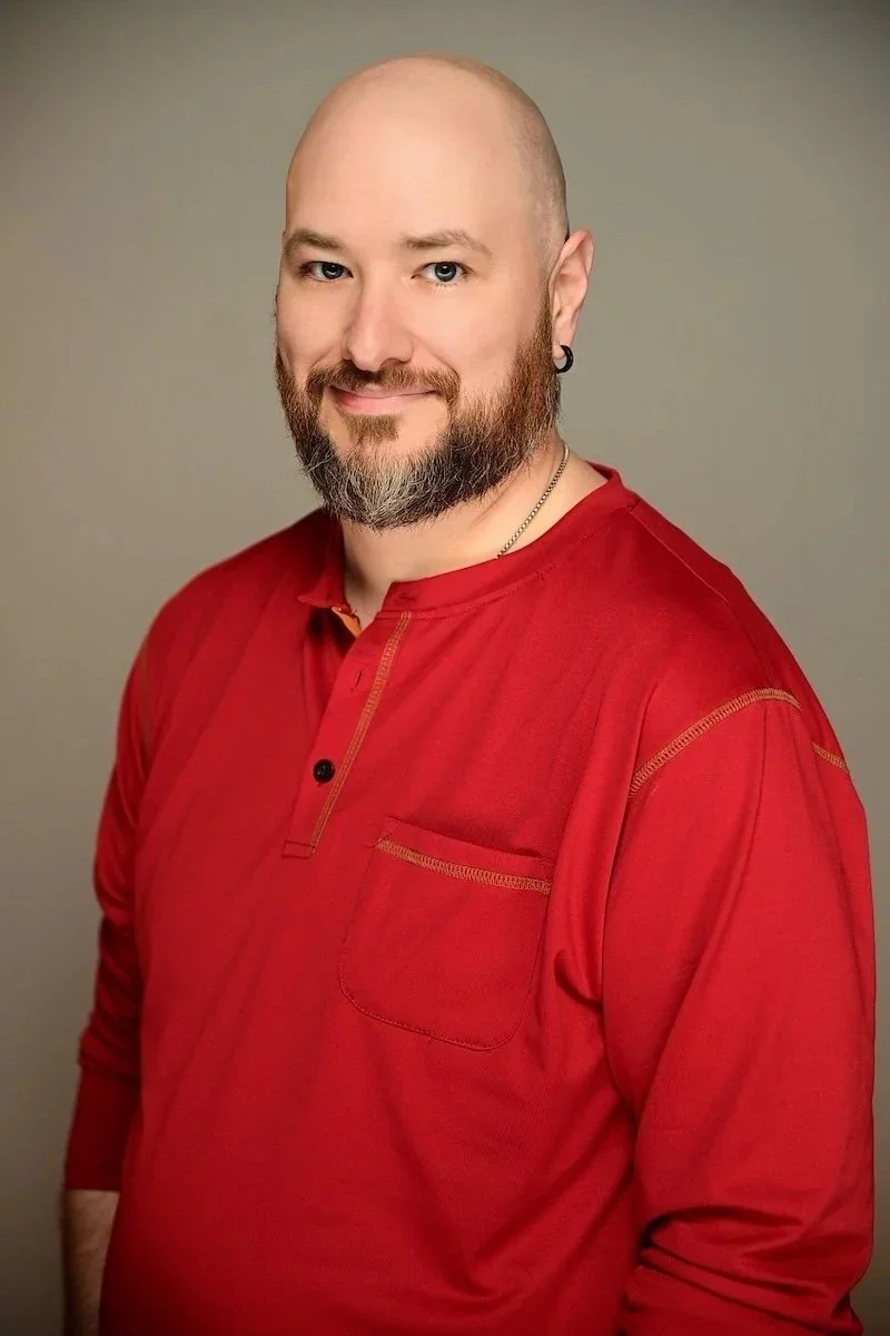 Smiling bald person with a short beard wearing a red long-sleeve shirt against a neutral gray background.