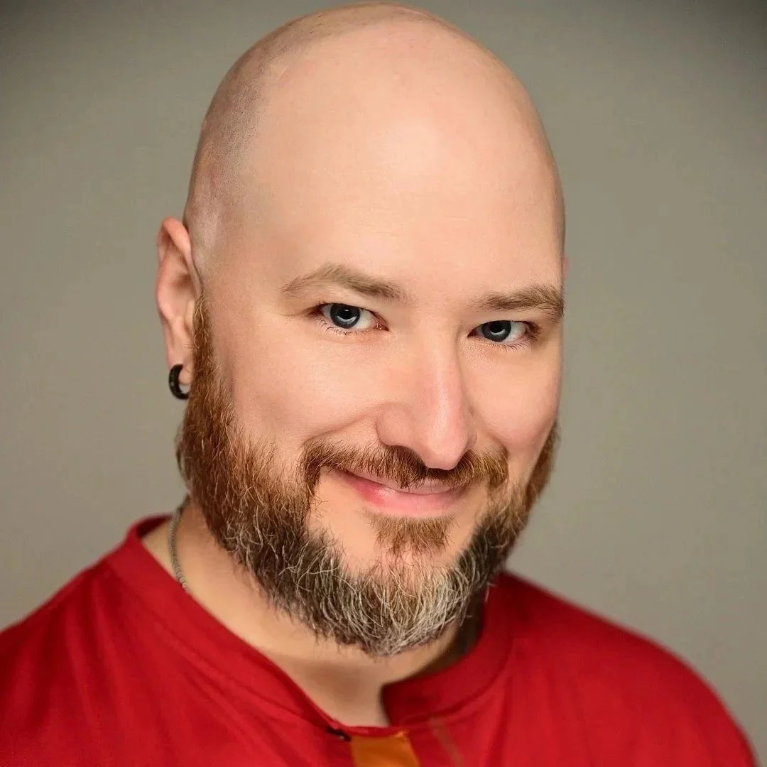 Professional headshot of a smiling bald person with a short salt-and-pepper beard, wearing a red henley shirt against a neutral gray background.