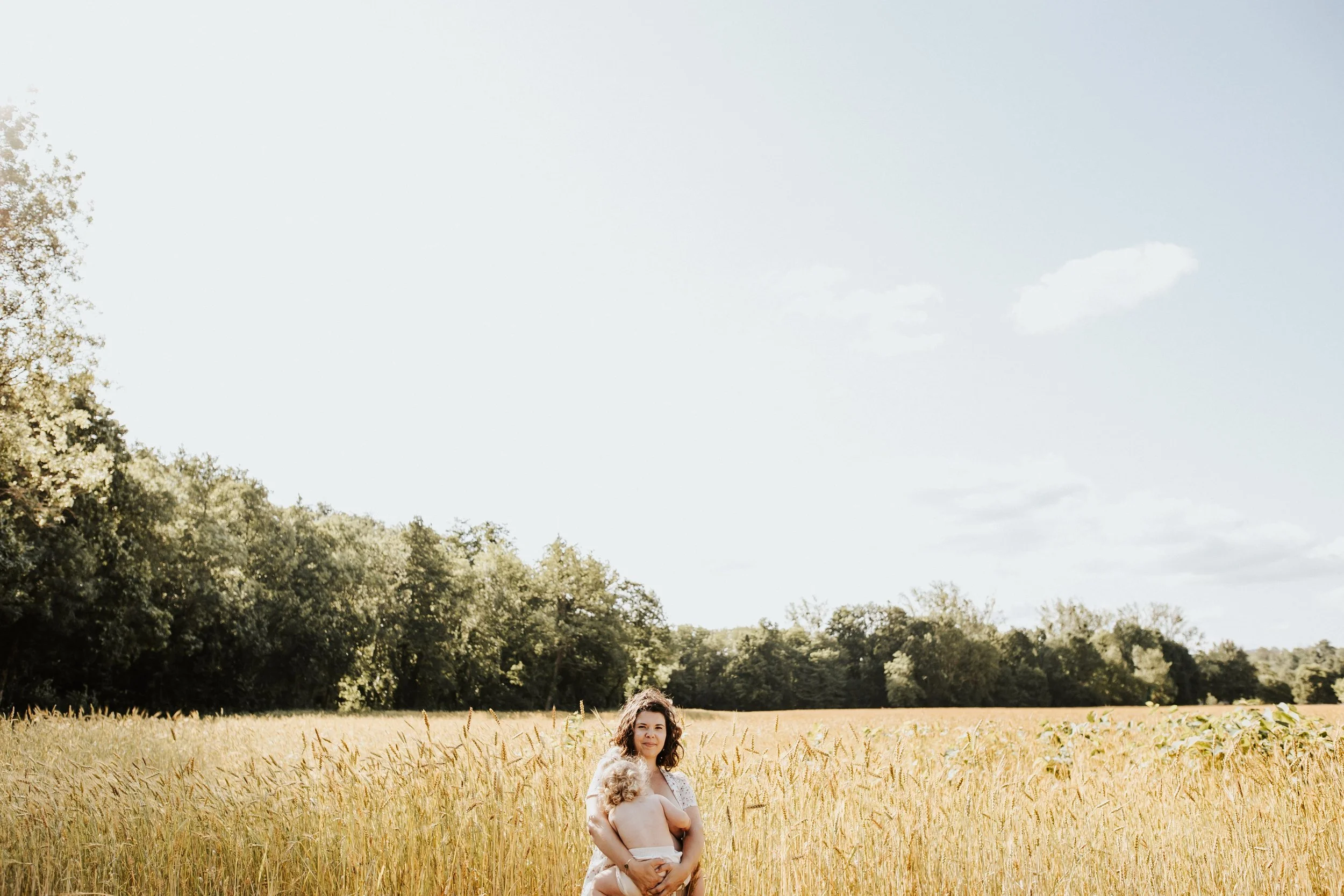 photo couleur d'une maman allaitant son enfant debout devant un champs de blé avec beaucoup d'espace négatif dans le ciel bleu du beaujolais près de Lyon