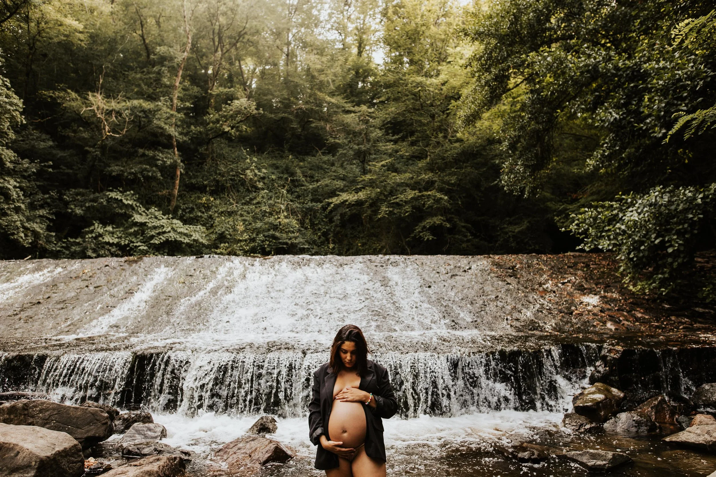 photo d'une future maman nue avec un blazer devant une cascade en pleine nature dans le beaujolais