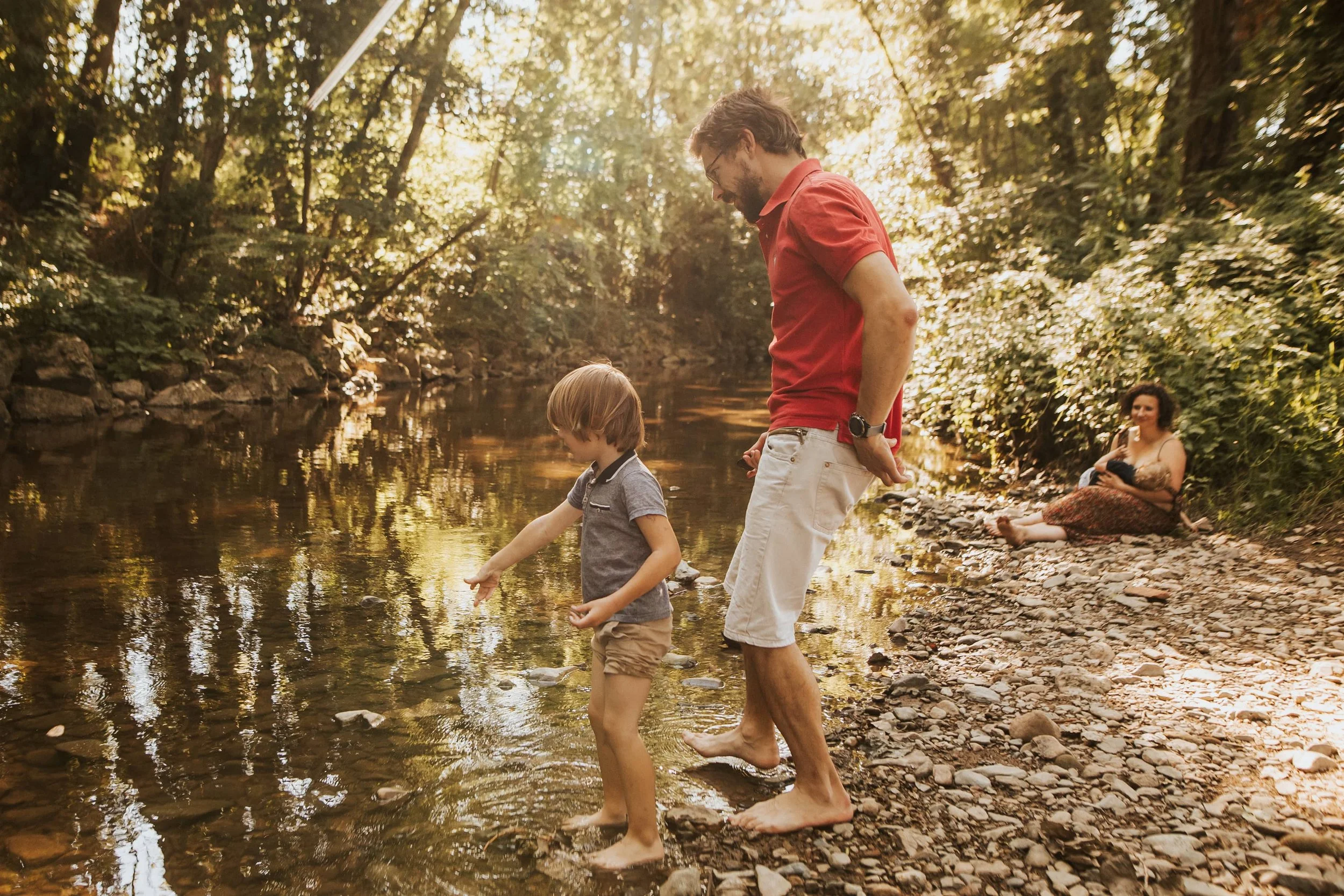 photo d'un papa jouant pieds nus au ricochets avec son grand garçon dans une rivière  au premier plan avec au second plan la maman allaitant le plus petit , tout ça pendant leur séance famille près de lyon