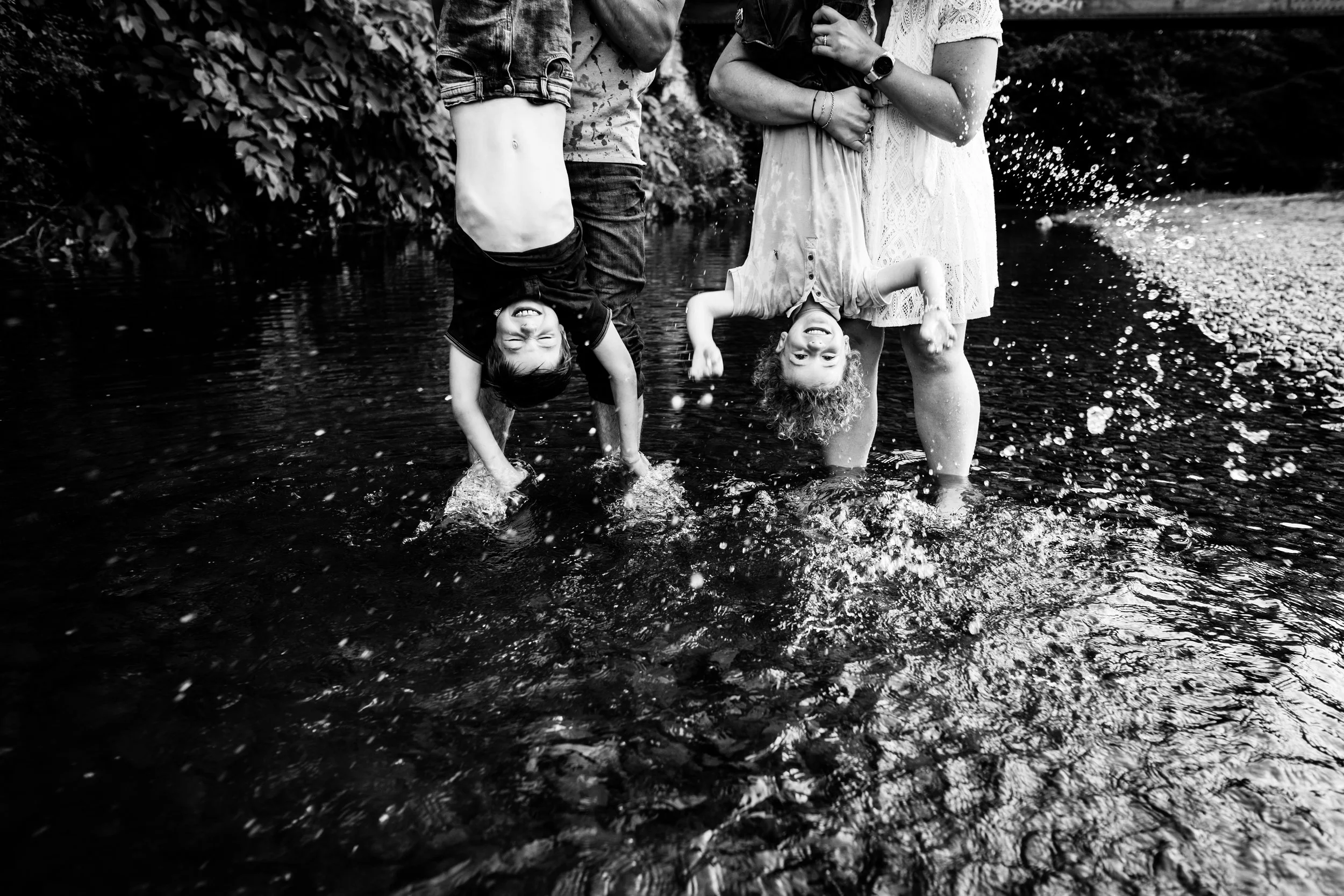 photo plein de joie noir et blanc de deux enfants la tête en bas tenus par leur parents au dessus d'une rivière, les mains qui éclaboussent, près de Lyon
