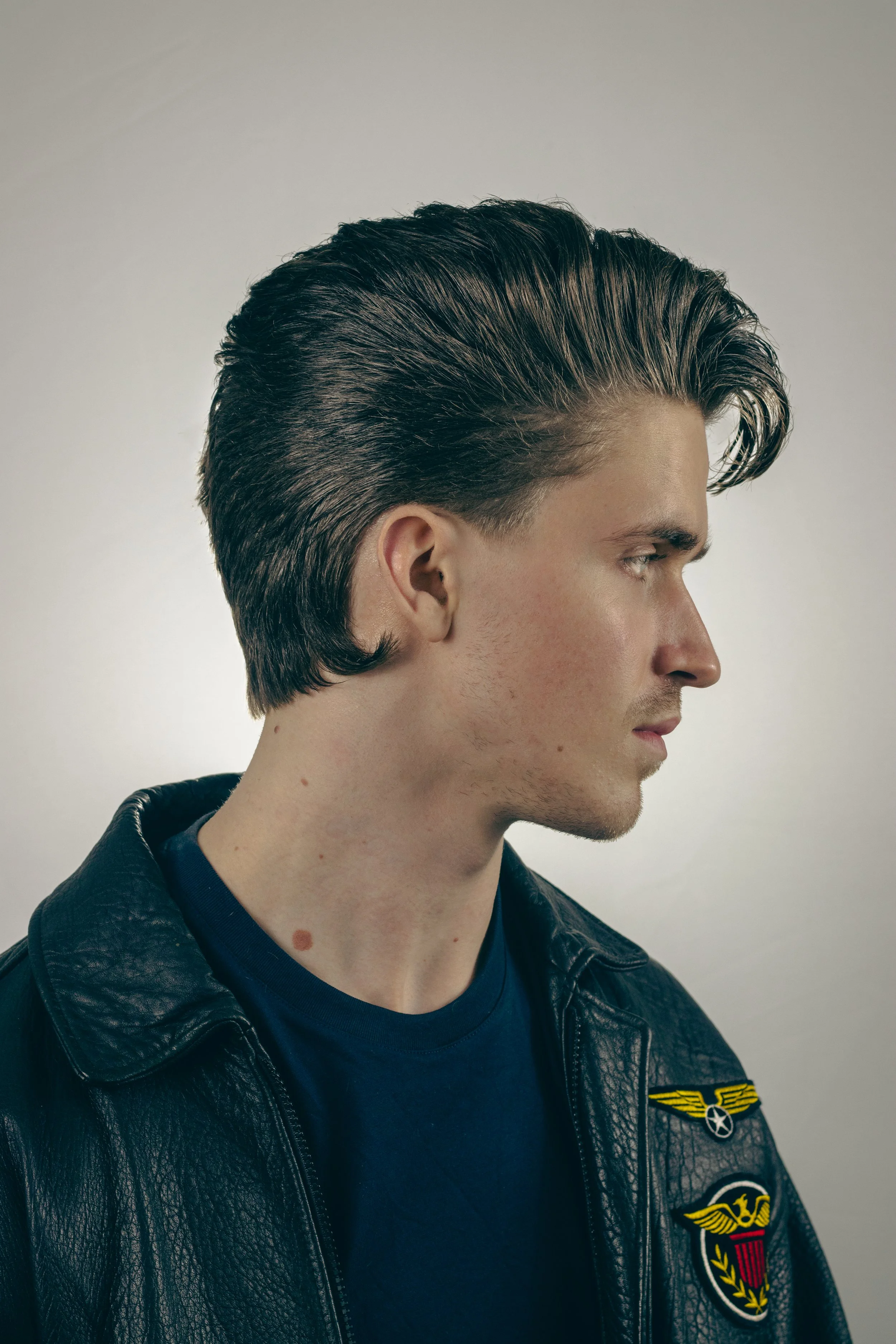 Side profile of a young man with dark, slicked-back hair, wearing a black leather jacket and a dark blue T-shirt, standing against a plain grey background.