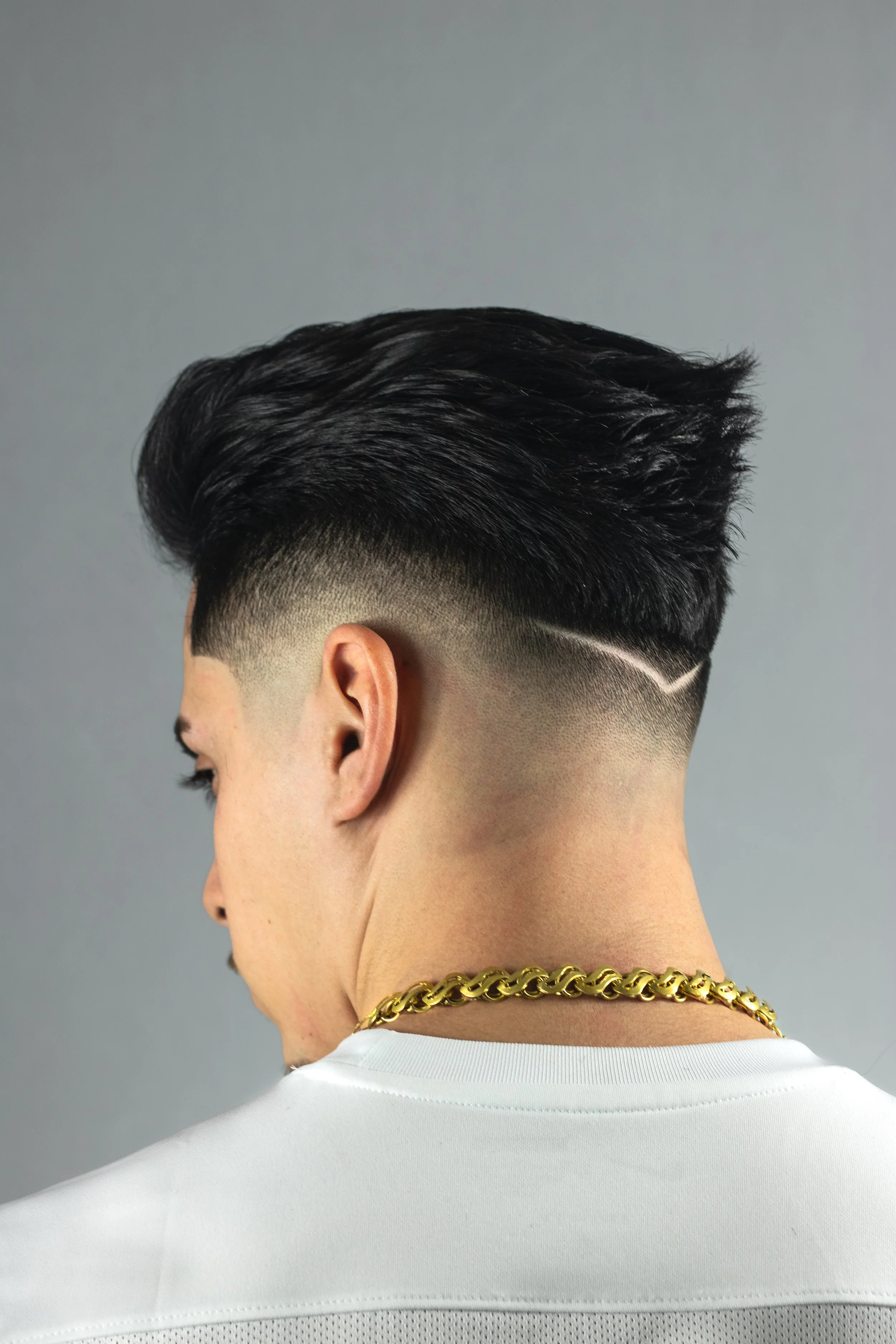 Close-up of a man's head with a modern haircut, featuring a fade and a white line design shaved into the side; he is wearing a white shirt and a gold chain necklace.