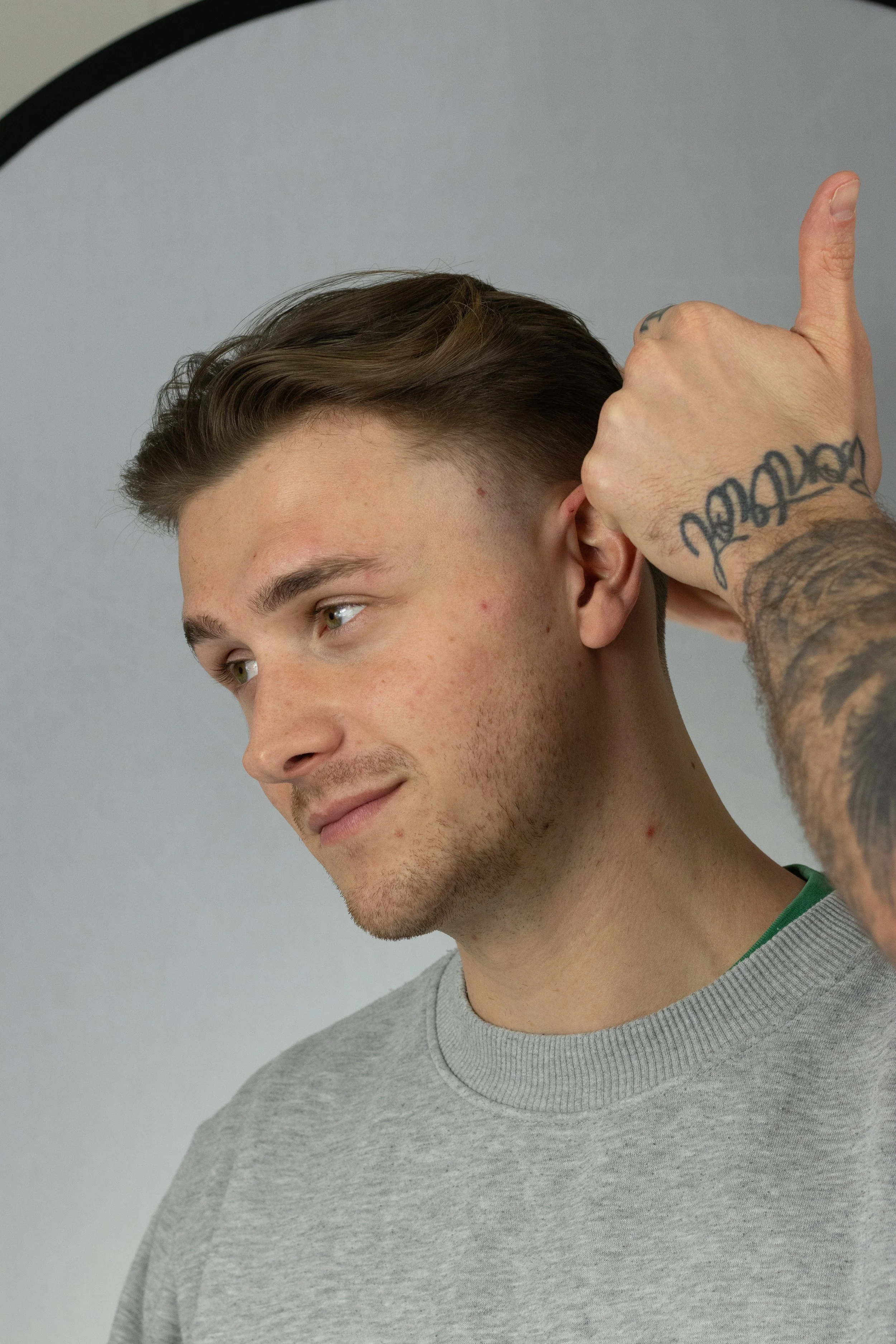 A young man with short brown hair and light stubble is getting his hair styled by a barber. The barber, who has tattoos on their arm, is giving a thumbs up and visible behind the man's right ear. The background is plain and grey.