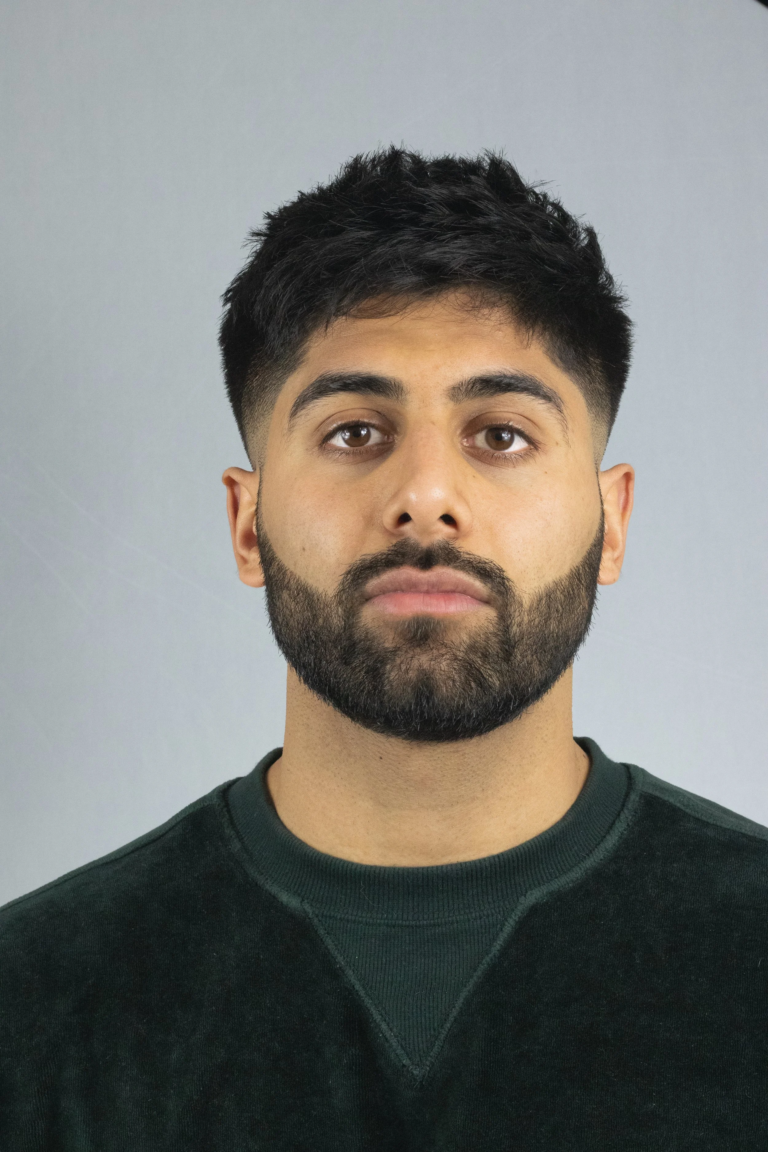 A man with short dark hair, a beard, and brown eyes looking directly at the camera, wearing a dark green shirt against a light gray background.