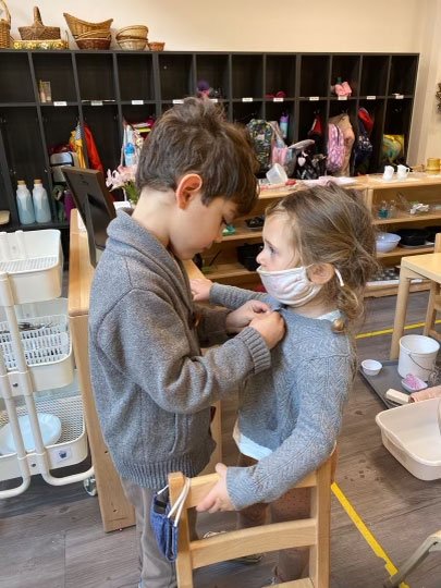  Kindergartners are the leaders of the Montessori classroom. Here in the Yellow Room, a third year student helps a first year student with her buttons while she looks on with admiration.  