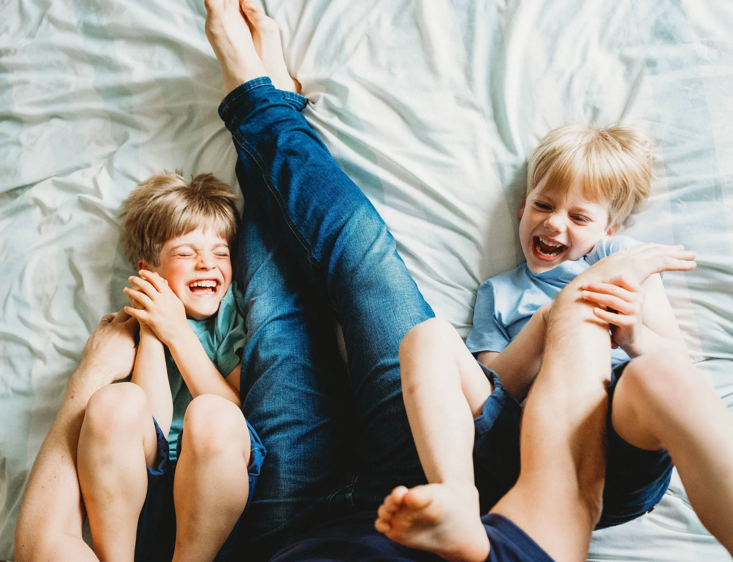 Two boys lie in a bed with their dad tickling them from above on a family photo shoot in Cheltenham