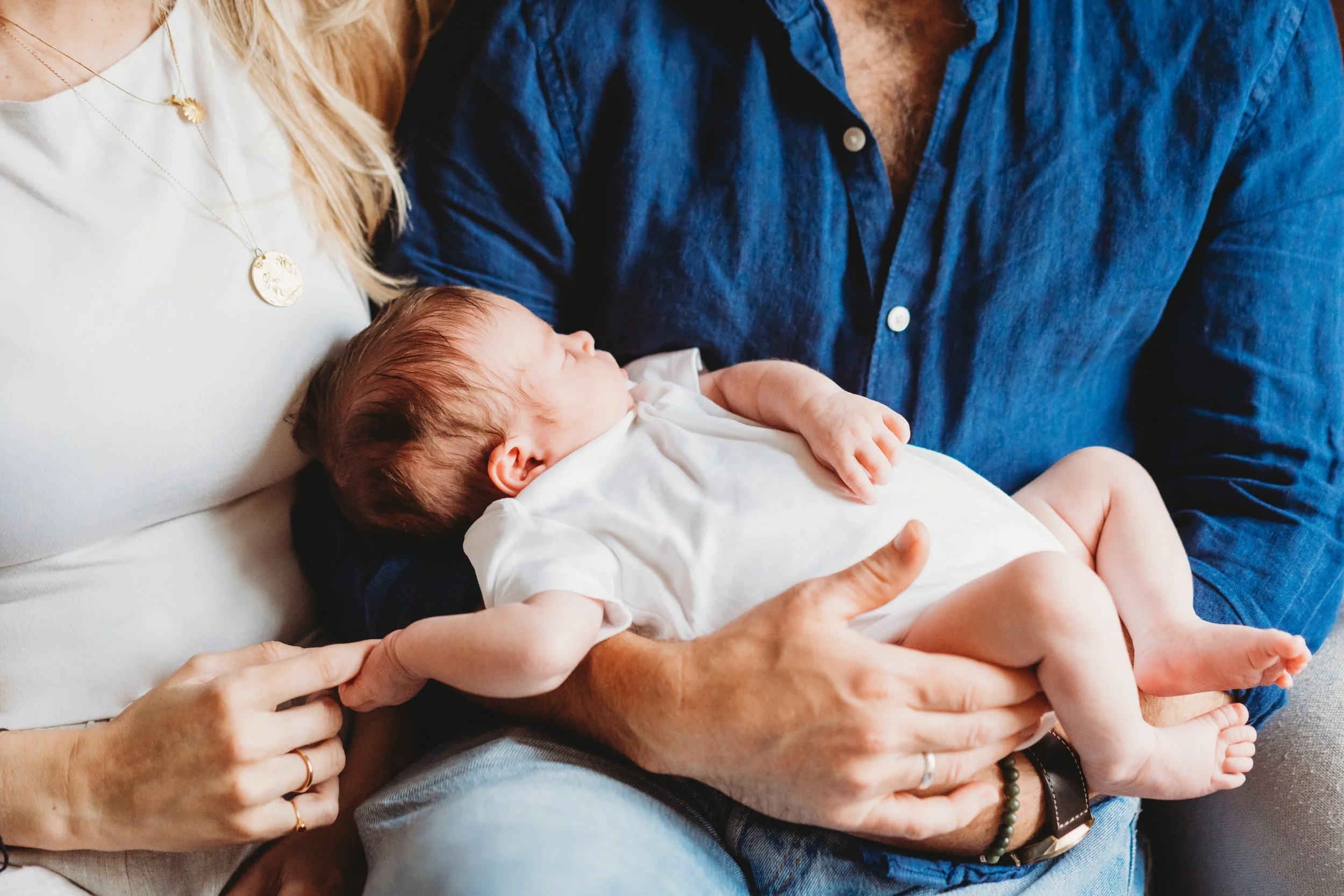 a newborn baby lies across her parents laps while the mum holds the baby's hand. The photo is cropped so you can't see the parents faces.