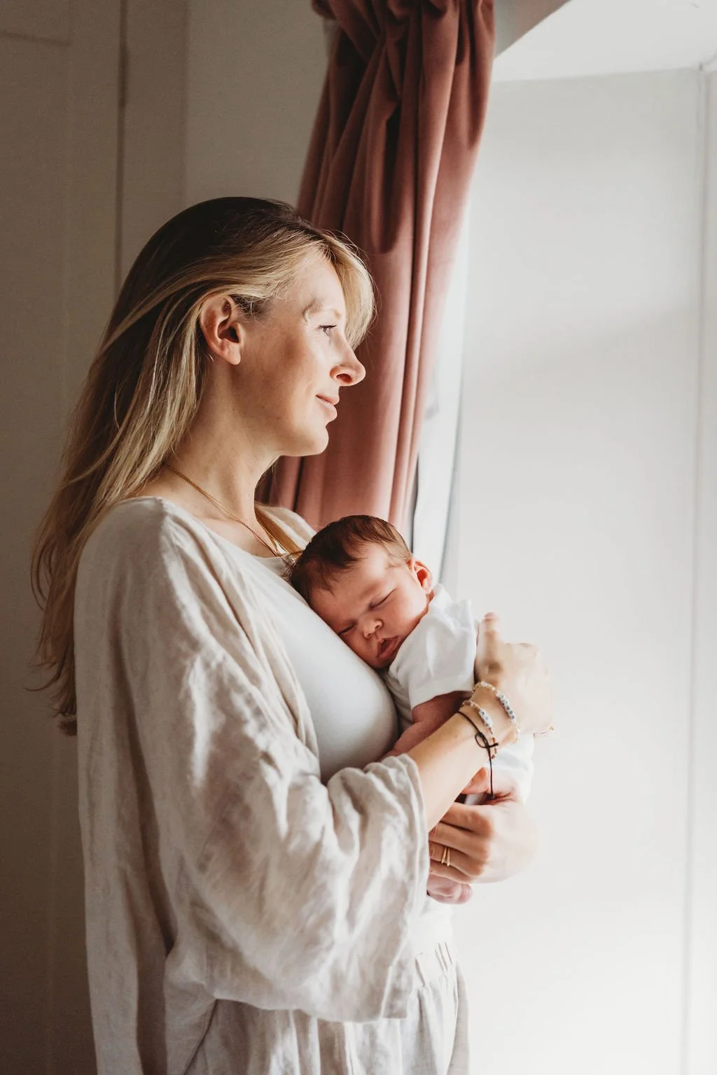 A new mum holds her newborn baby against her chest while looking out of the window she is stood next to during a newborn baby photoshoot in Cheltenham