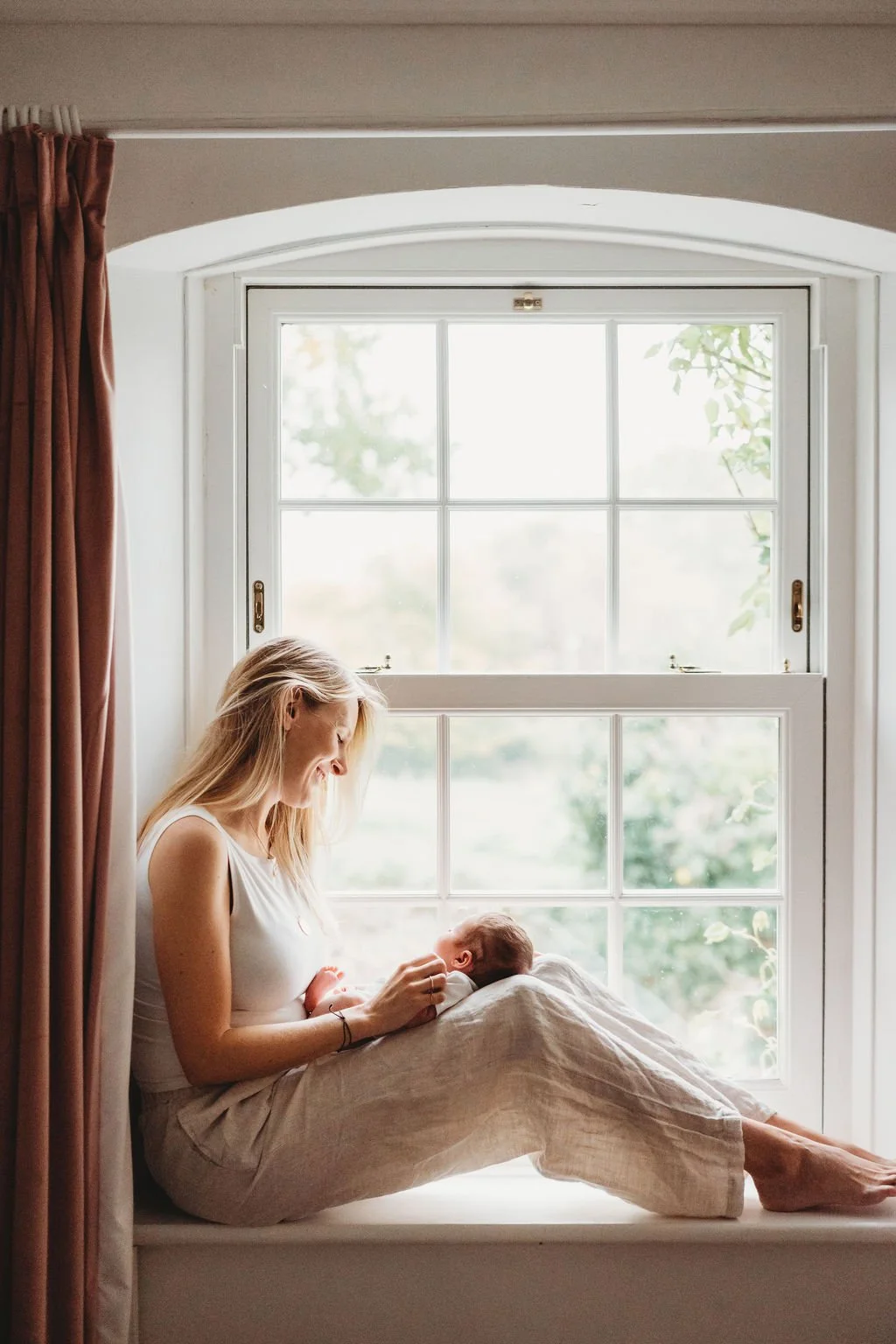 A mother sits on the window sill of a large Georgian style window with her knees up in front of her, parallel to the photographer.  Lying on her knees is her newborn baby who is asleep during a newborn baby photo shoot in Cheltenham