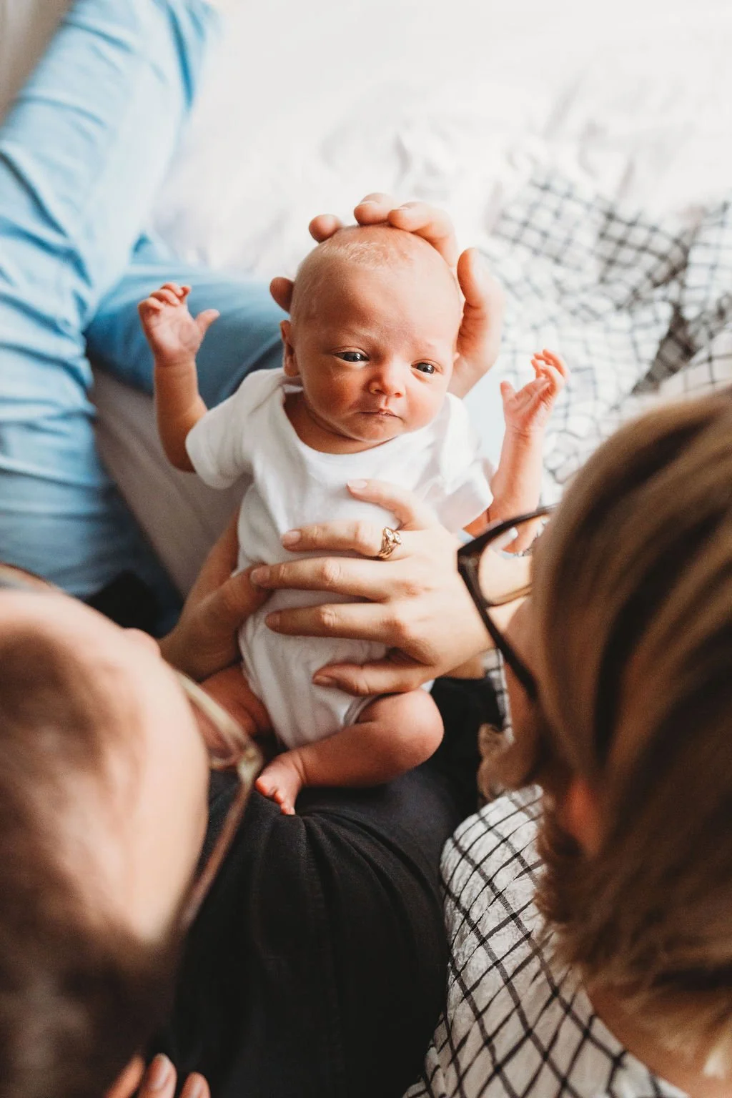 A newborn baby lies in her parents arms looking up at them while the photographer photographs over the parents shoulders so the shot is looking down onto the baby during a newborn baby photoshoot in Cheltenham