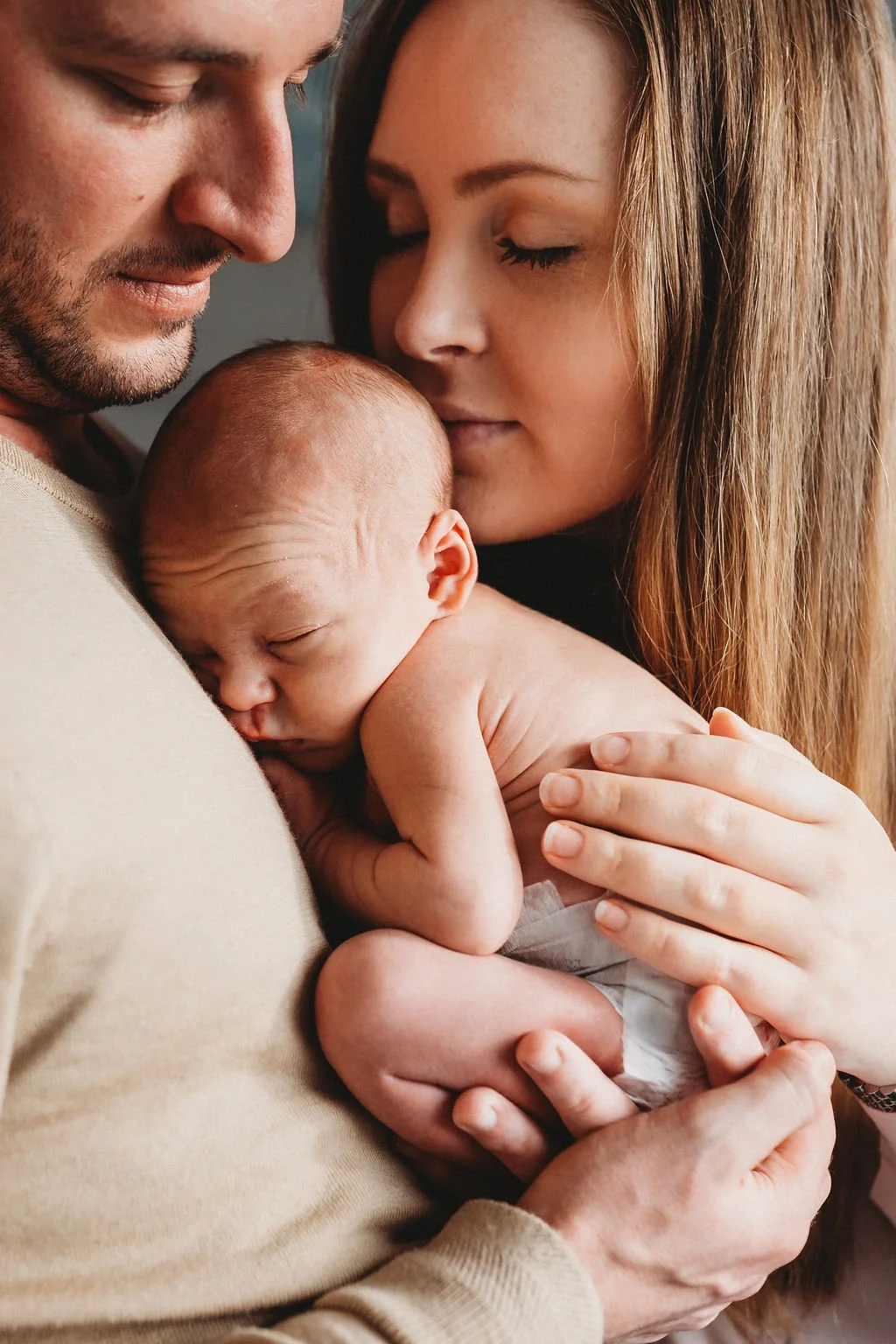 A newborn baby lies against his father's chest while his mum leans to kiss him on the back of his head during a newborn photo shoot in Cheltenham
