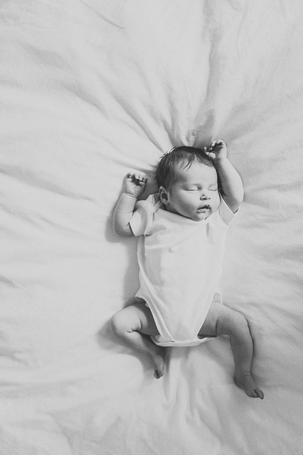 A newborn baby lies asleep on a bed in a white vest. The photo is taken from above and is in black and white during a newborn baby photo shoot in Cheltenham