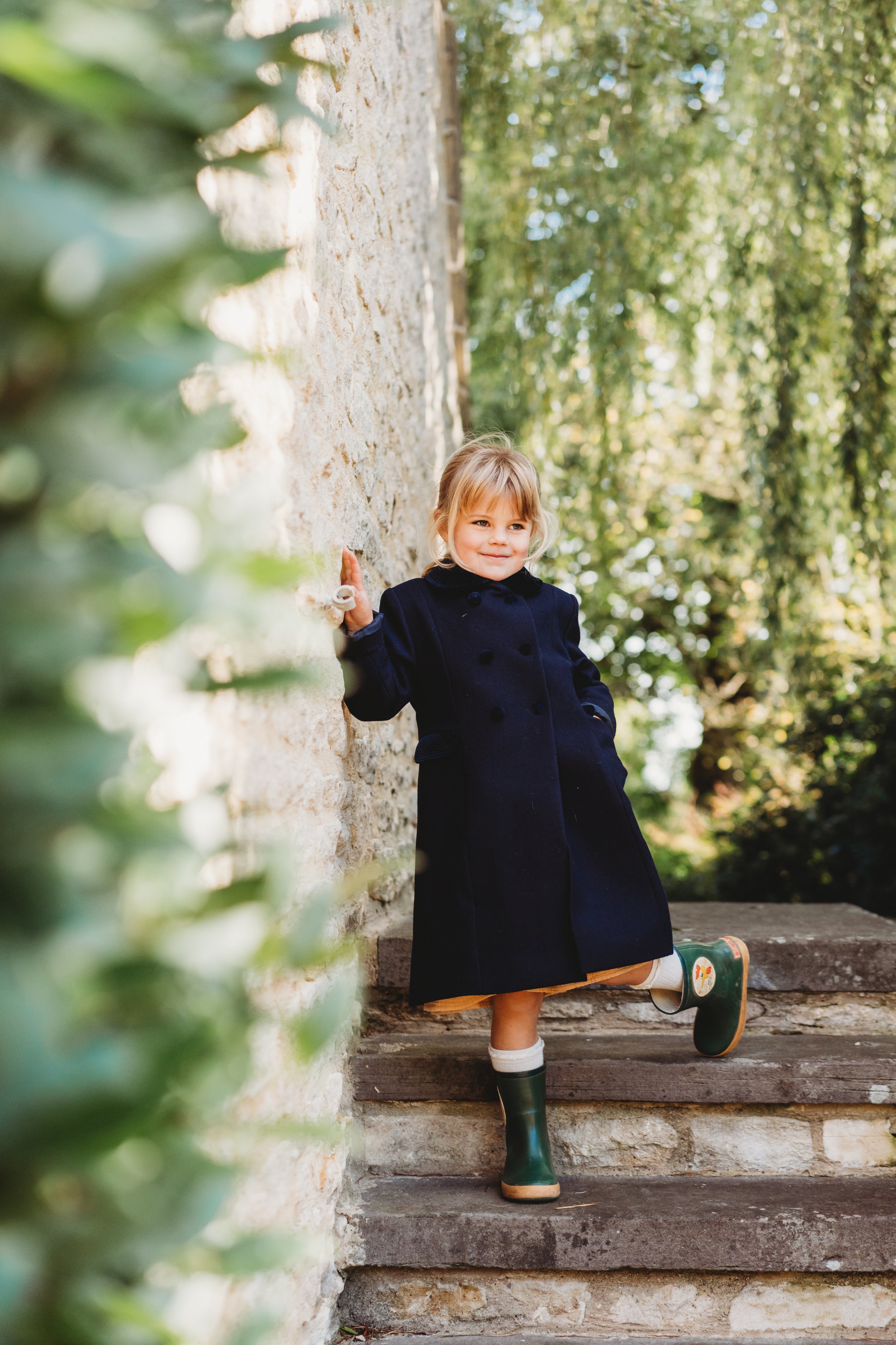 A little girl stands on steps next to a Cotswold stone barn.  She has one hand in her pocket and looks over the photographers shoulder smiling during a family photo shoot in The Cotswolds with Claire Westaway Photography
