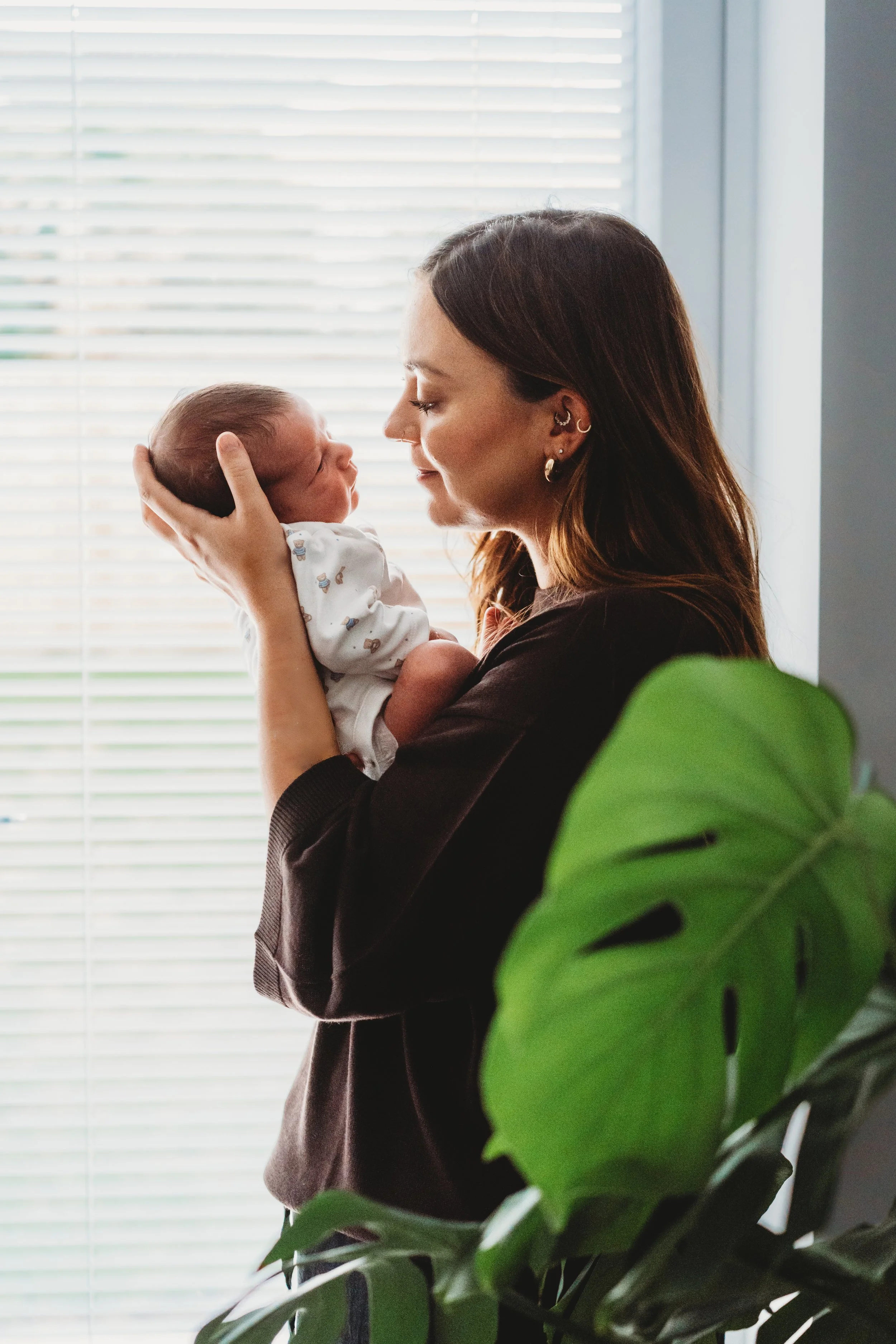 A new mum stands in front of a window cradling her newborn baby and they look at each other with their noses almost touching during a newborn baby photoshoot in Cheltenham.