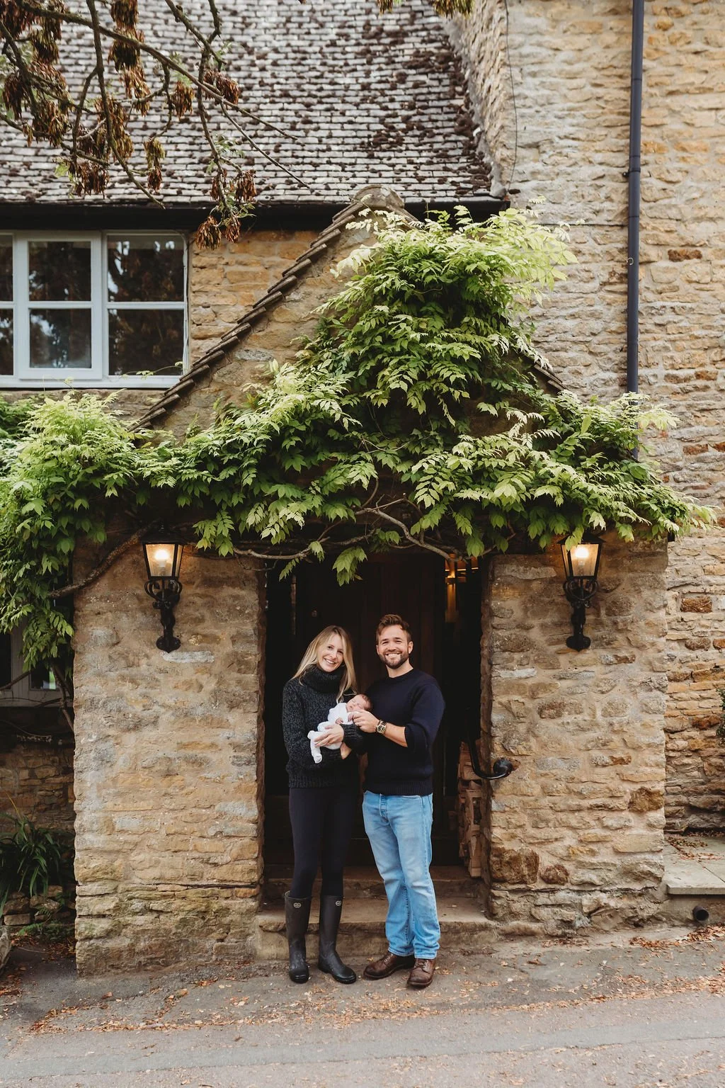 A young couple with their newborn baby stand outside the front door of a Cotswold pub in their village during a newborn baby photoshoot in Cheltenham