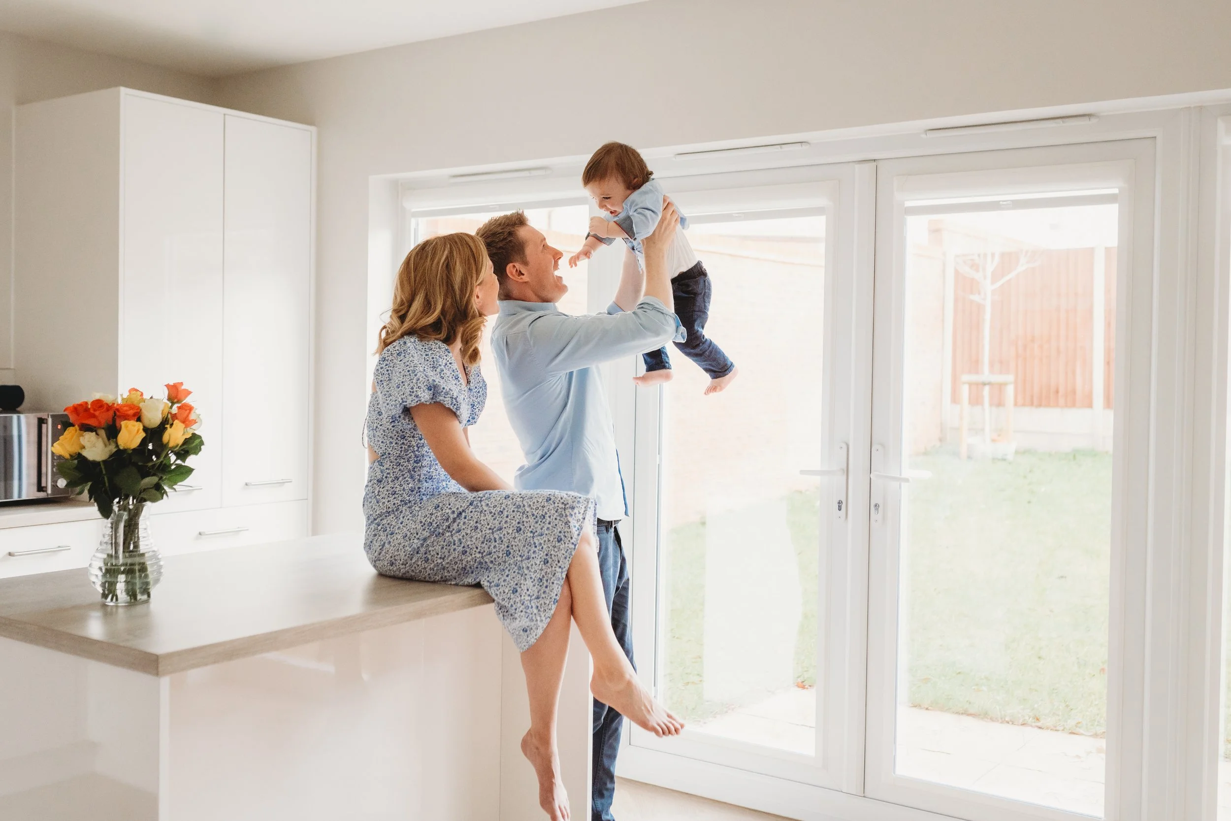 In a light white kitchen, a mum sits on the island in a blue dress, a dad stands next to her holding their baby up in the air. The parents are both looking at the baby during a family photo shoot in Cheltenham.