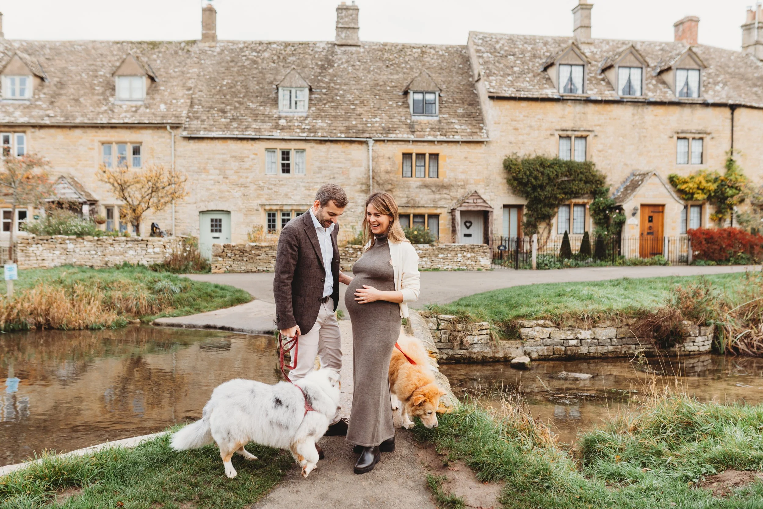 in the village of Lower Slaughter, a pregnant woman stands next to her husband and their two dogs on a bridge going over a stream.  The Cotswold cottages are in the background during a Maternity photo shoot in The Cotswolds.