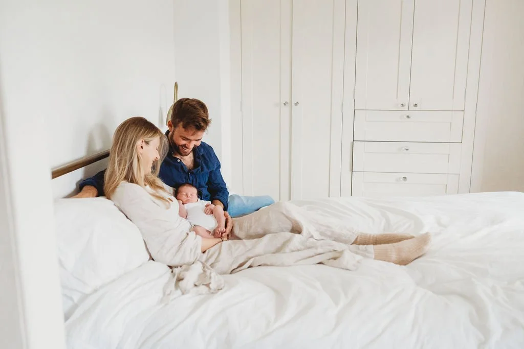 A young couple sit on their bed holding their newborn baby in their arms who is fast asleep during a newborn baby photo shoot in Cheltenham.