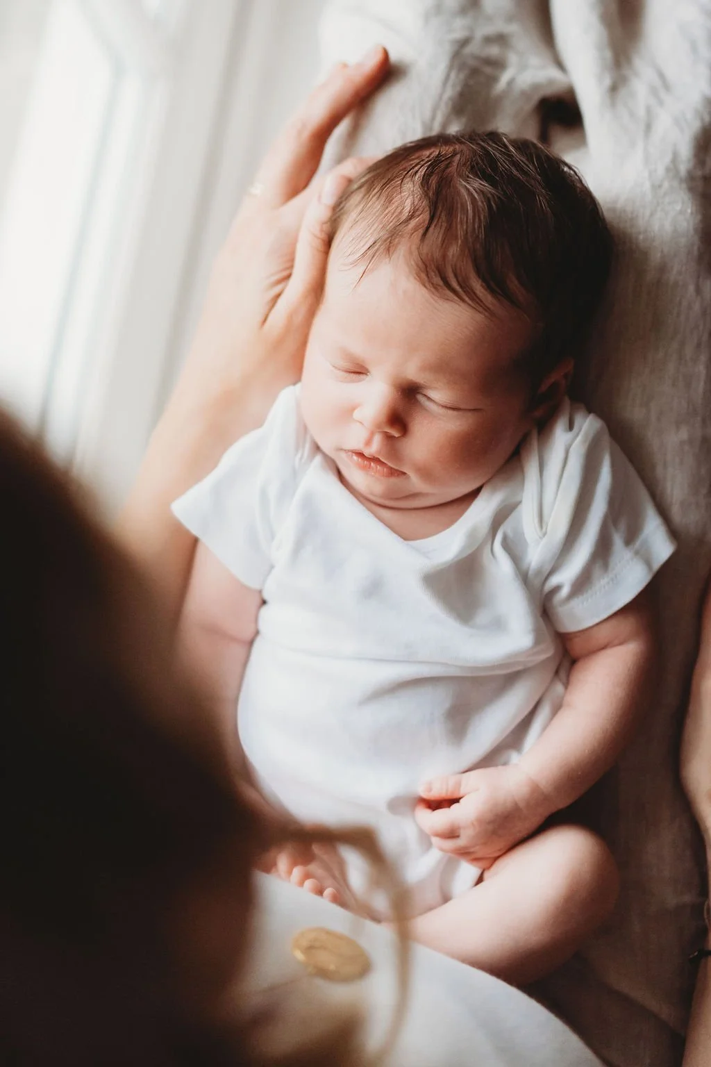 a newborn baby lies on her mother's lap while the photographer takes the shot from over the mum's shoulder so you are looking down on her asleep during a newborn baby photoshoot in Cheltenham