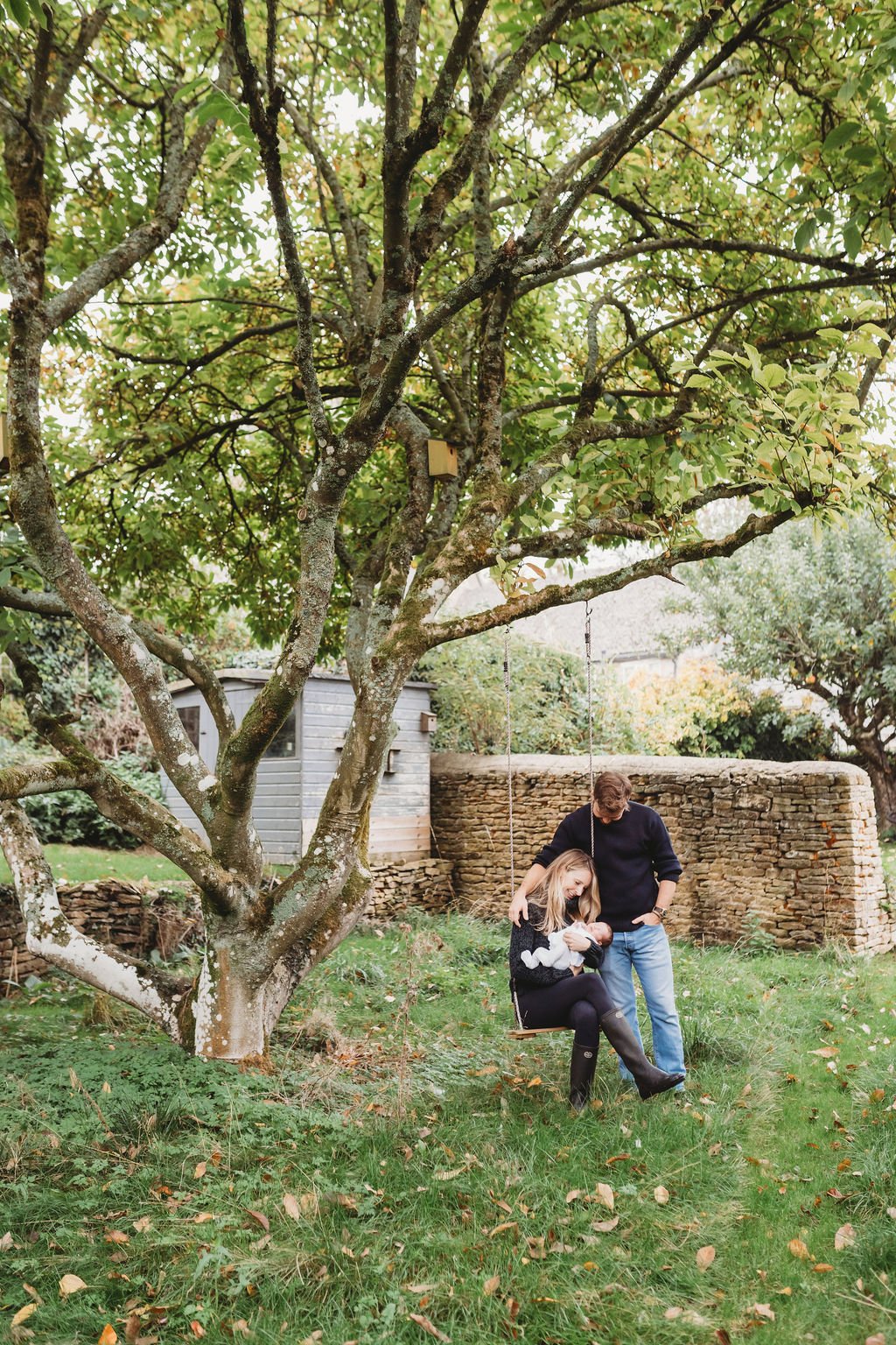 In a garden, a new mum sits on a swing hanging from a tree craadling her newborn baby while the father stands alongside during a baby photoshoot in Cheltenham.