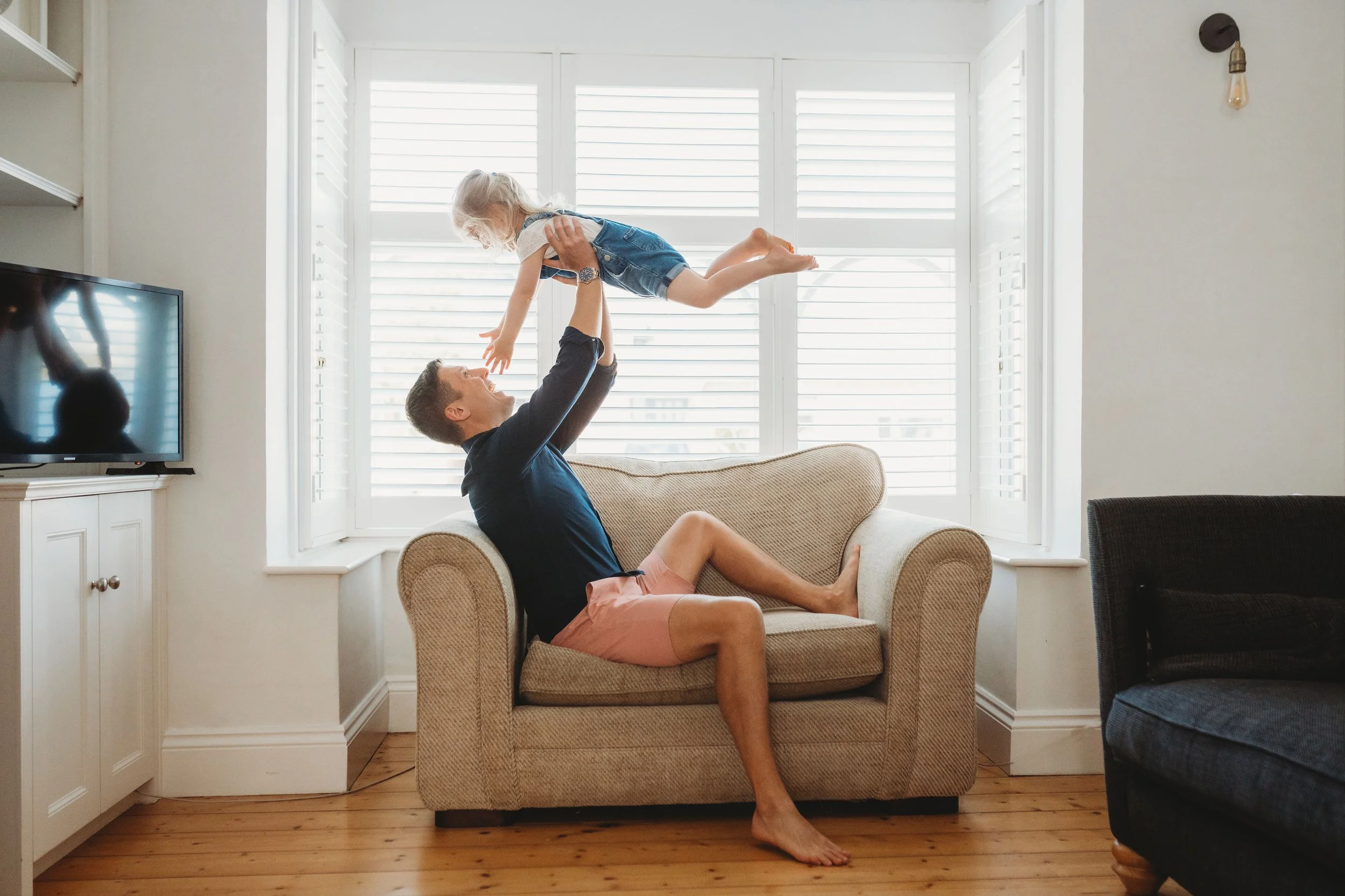 A dad is sat on a chair in front of a large window in the lounge and he lifts his young daughter up in the air.  Her hands reach down to try and touch his face during a family photo shoot in Cheltenham.