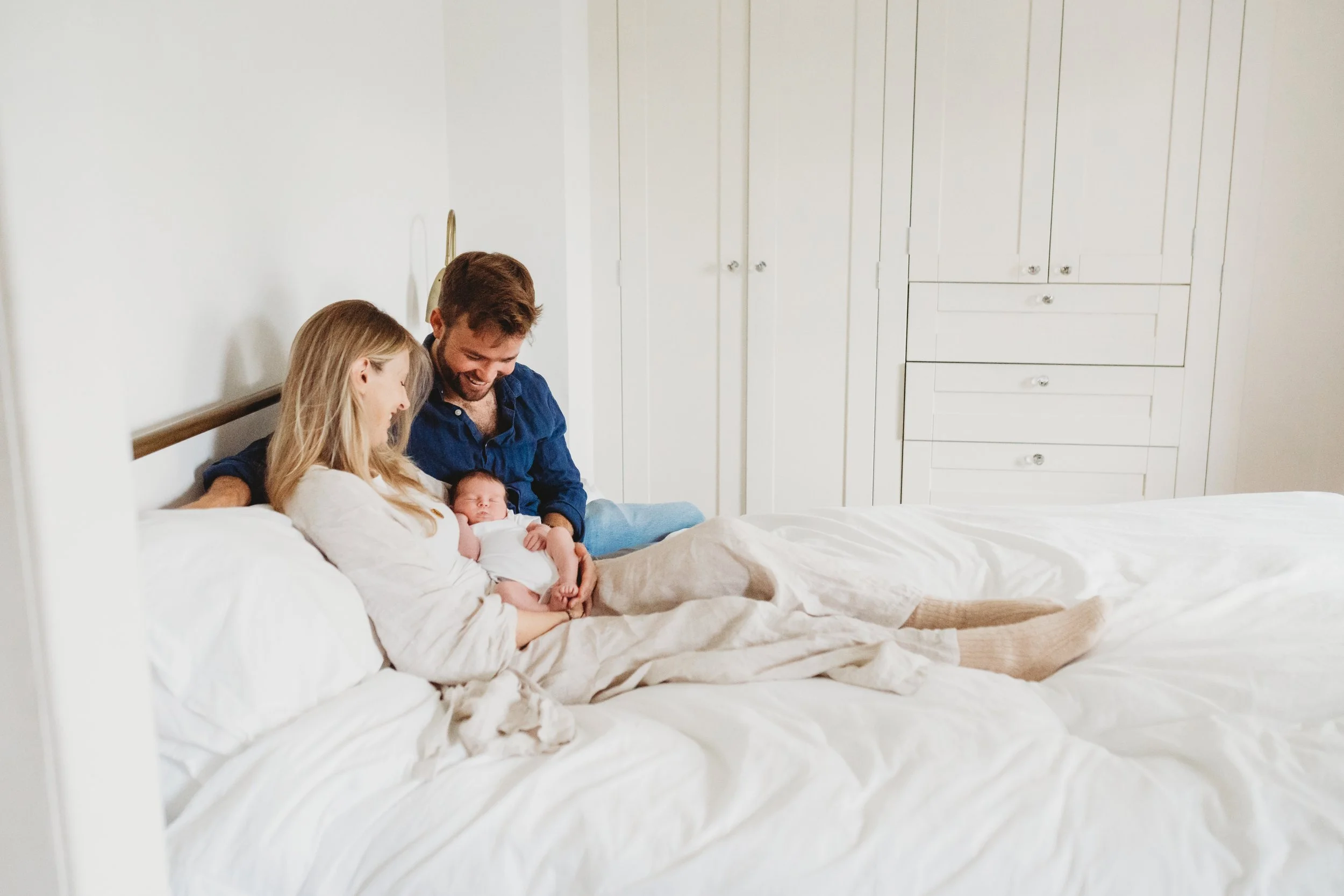 A new mother and father sit on their bed looking down at the newborn baby in their arms during a newborn baby photo shoot in the cotswolds.