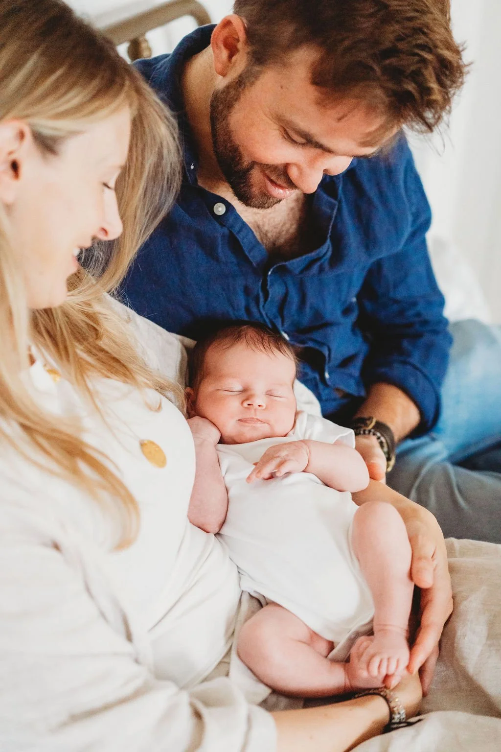 a newborn baby lies between her parents in their arms as they look down at her during a newborn baby photoshoot in Cheltenham