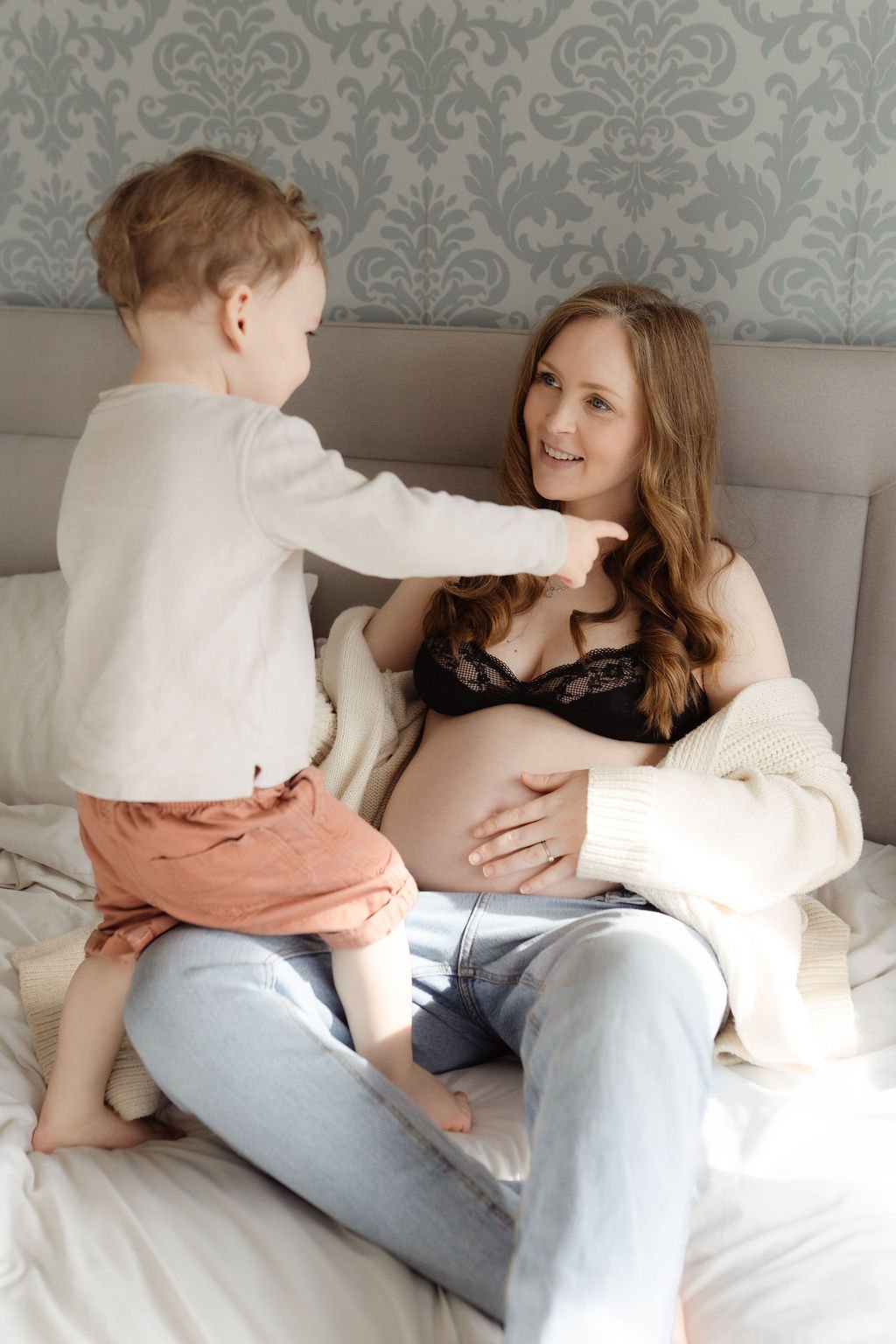 A pregnant mum sits on a bed with her bump showing while her toddler stands in front of her and they are both laughing during a maternity photo shoot in Cheltenham, The Cotswolds.