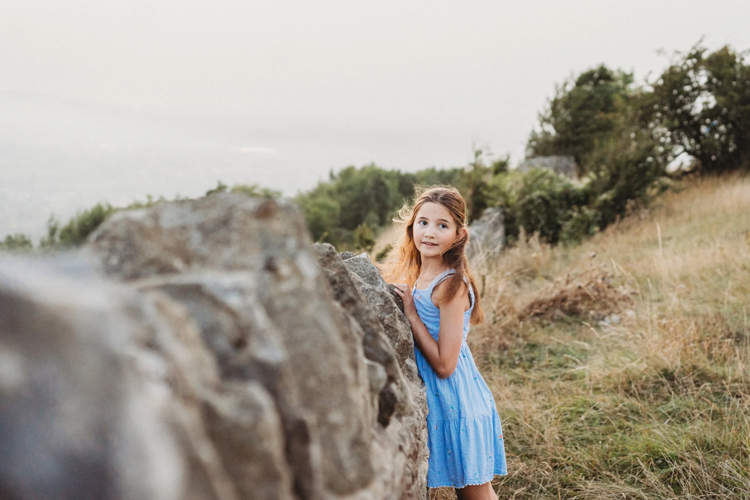 a young girl leans on a Cotswold stone wall and looks over her shoulder with the wind blowing her hair whilst on a family photo shoot in Cheltenham.
