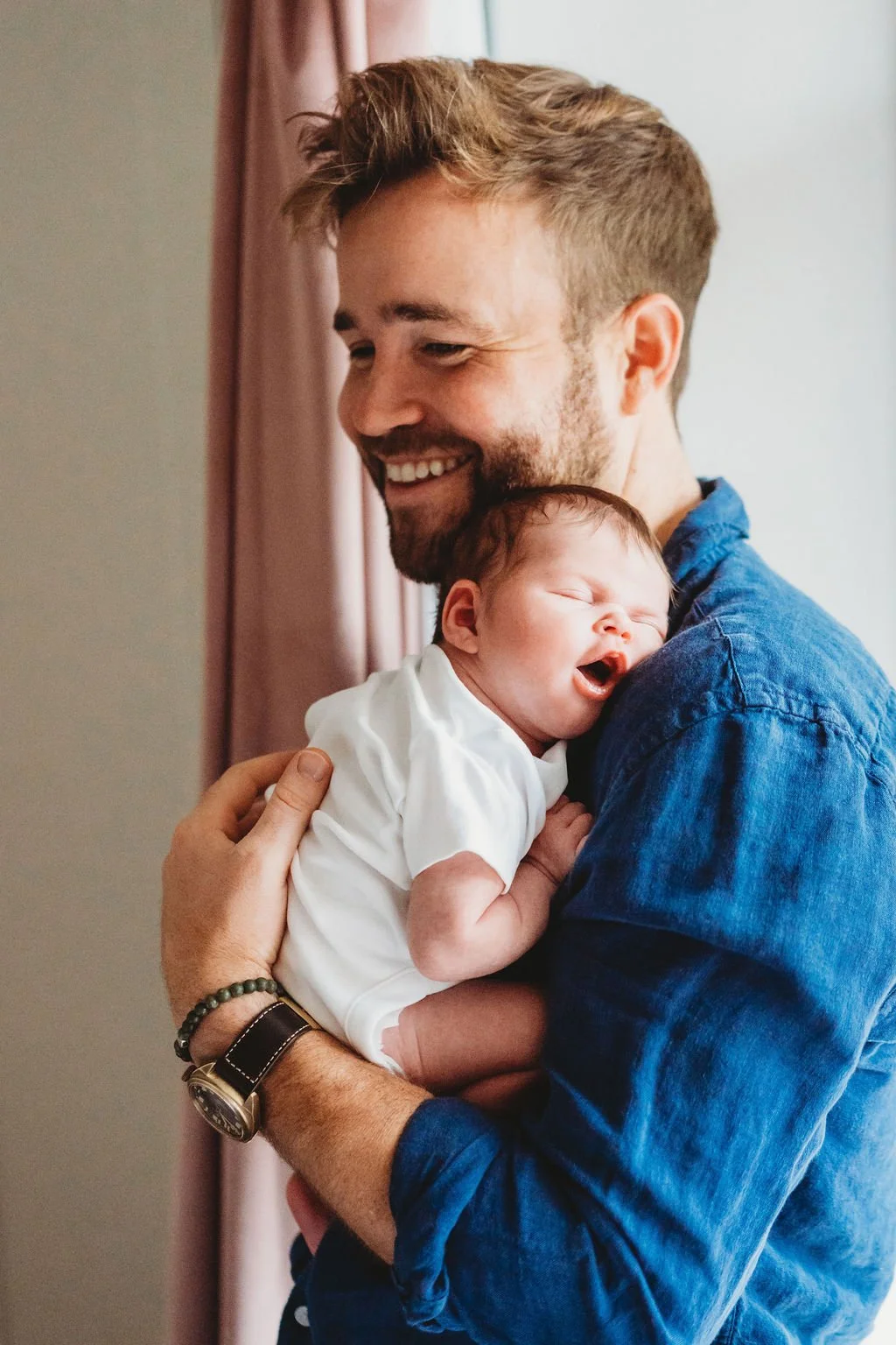 A father in a blue shirt holds his newborn baby against his chest.  The baby is asleep with her mouth open and the Dad is smiling during a newborn baby photo shoot in THE COTSWOLDS