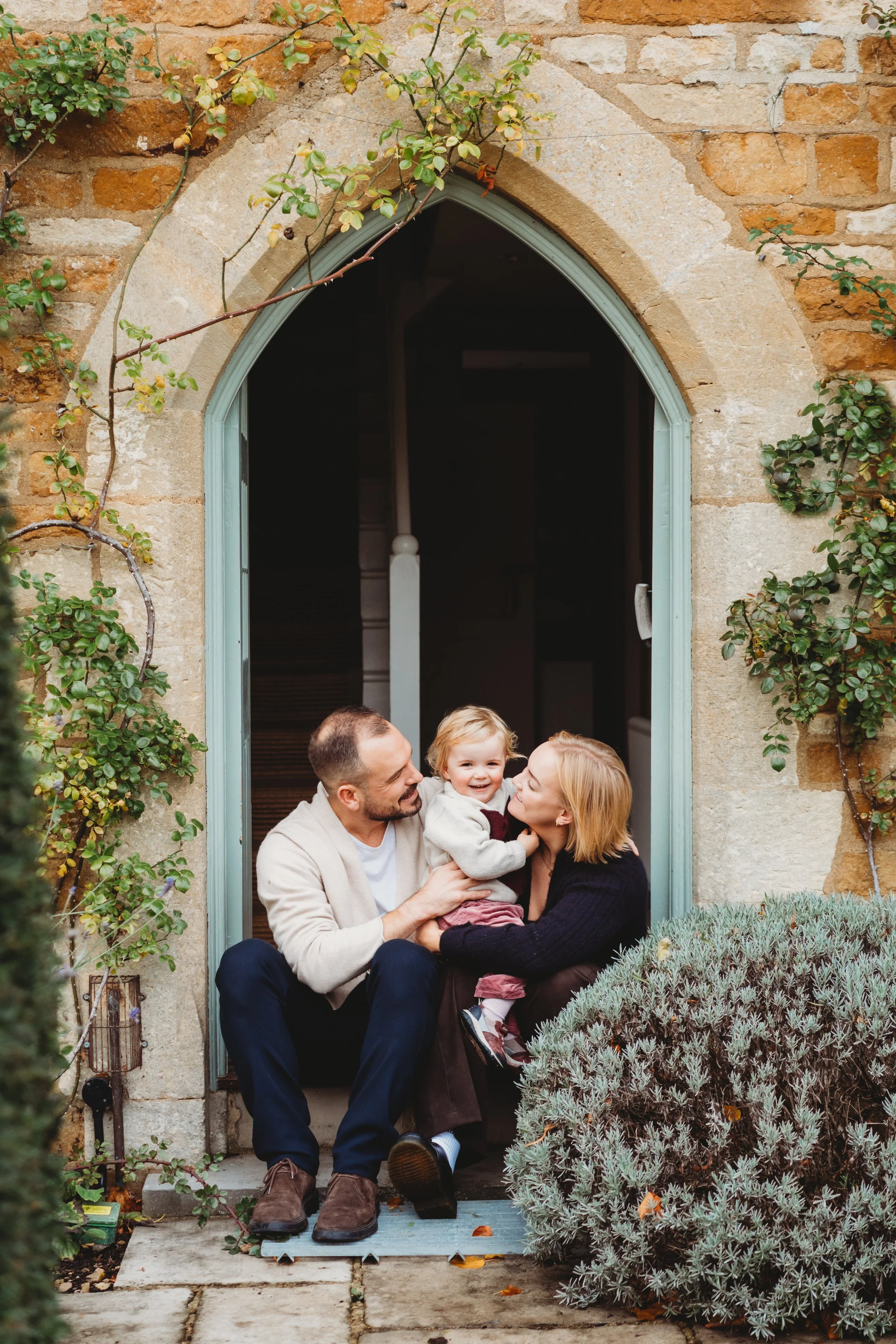 A family of mum, dad and toddler daughter sit on the step of their front door.  The parents are both looking at their daughter who is in the middle of them smiling at the camera during a family photo shoot in Cheltenham.