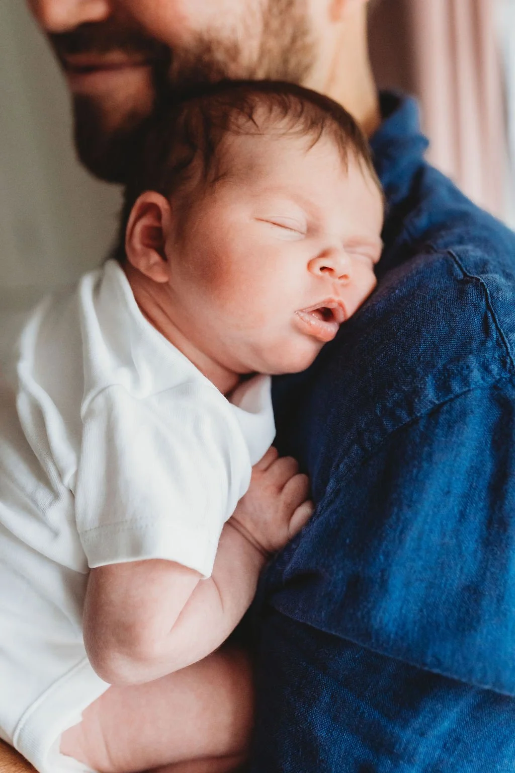 The Photo is a close up of a newborn baby who is fast asleep against her dad's chest and shoulder.  You can just see Dad's head behind the baby and he is smiling during a newborn baby photoshoot in Cheltenham