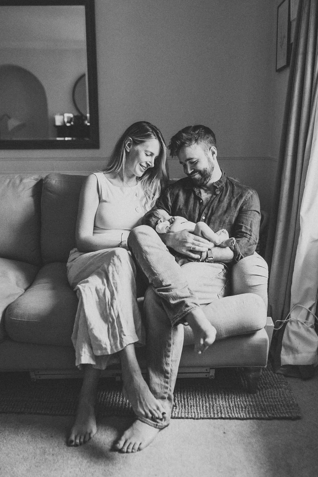 a new mum and dad sit on their sofa next to a window holding their newborn baby in their arms and looking down at her during a baby photoshoot in Cheltenham