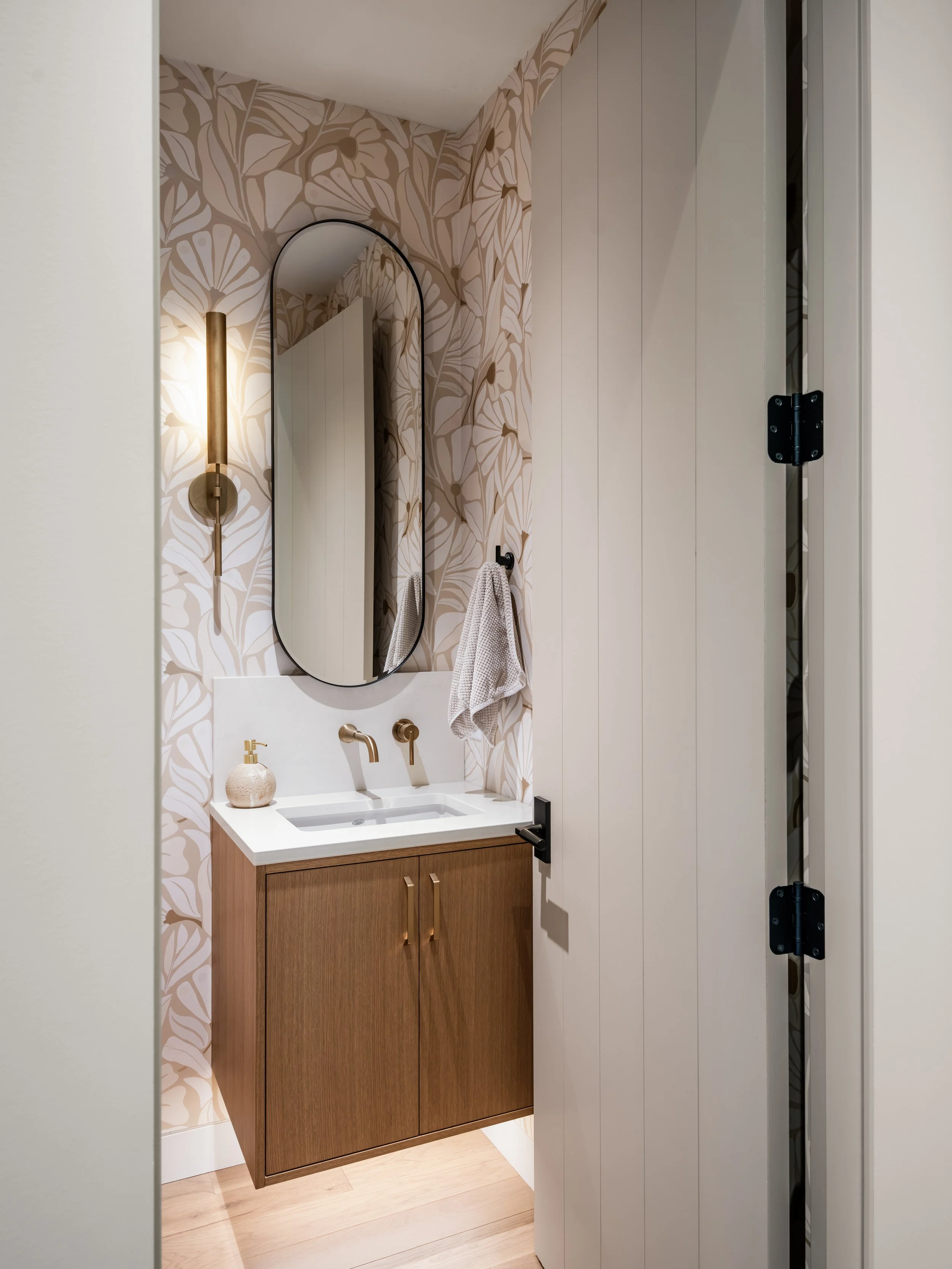 A small bathroom vanity with a modern white sink, gold faucet, and wooden cabinet, with a wall-mounted gold and white soap dispenser and a beige hand towel hanging on a hook. The background features a floral wallpaper with beige and white tones, and an arched mirror above the vanity. To the right, there is a white panel wall with black hinges.