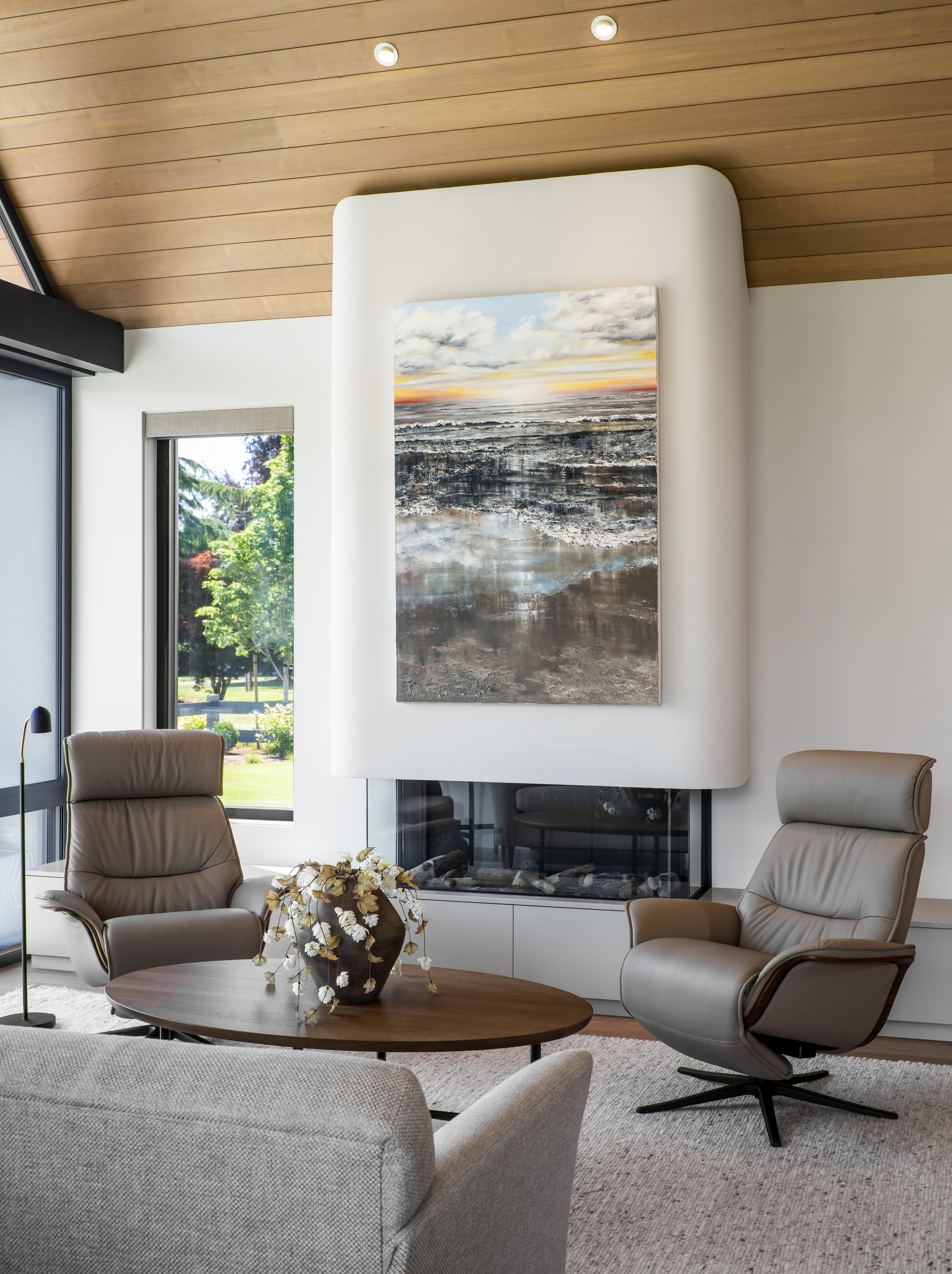 Modern living room with beige leather chairs around a round wooden coffee table with a black vase and flowers, large window showing green trees, and a white fireplace with a landscape painting above it, all under a wooden ceiling.