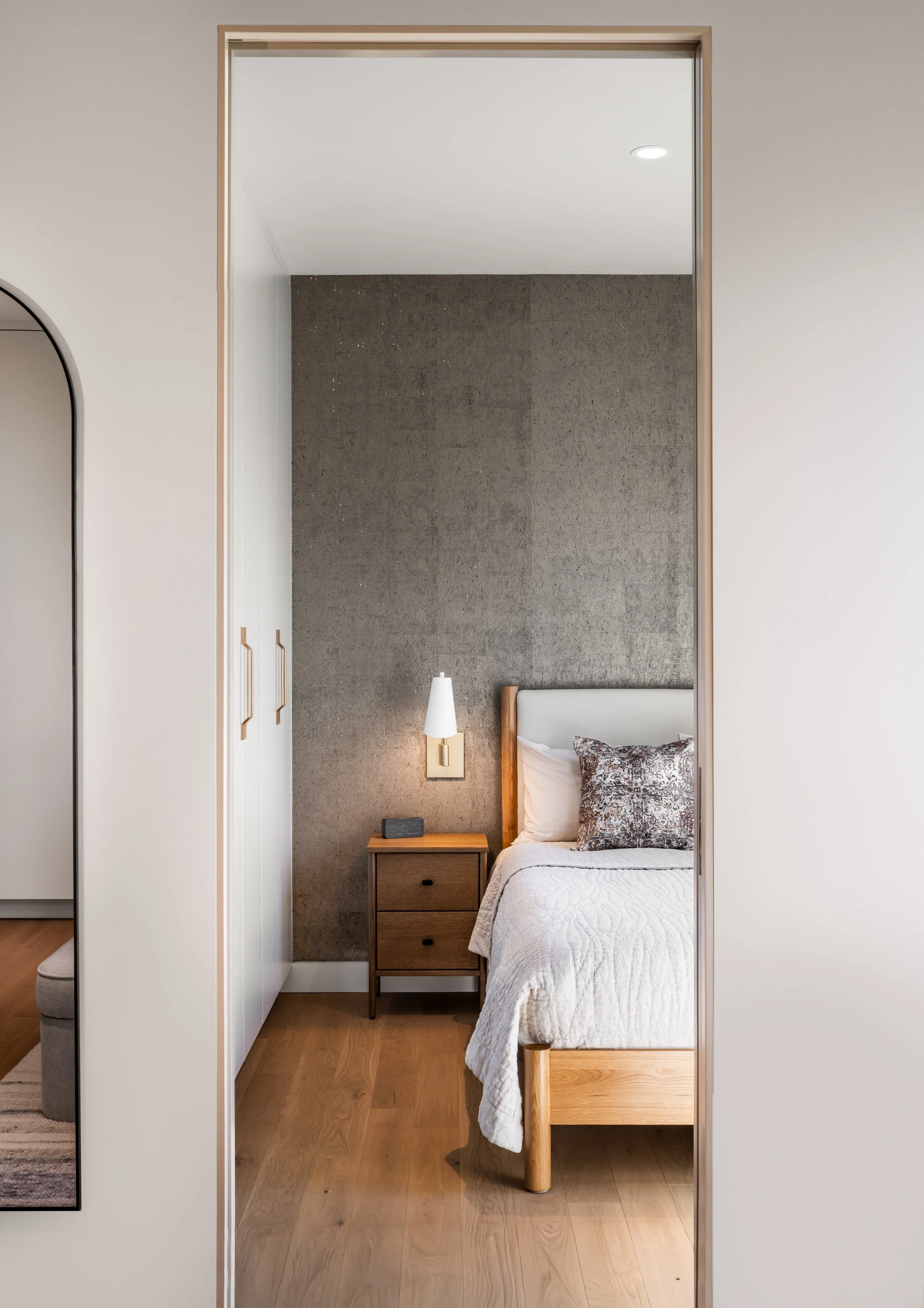 A bedroom with a bed, nightstand, and wall-mounted light, seen through an open doorway with a wood frame.