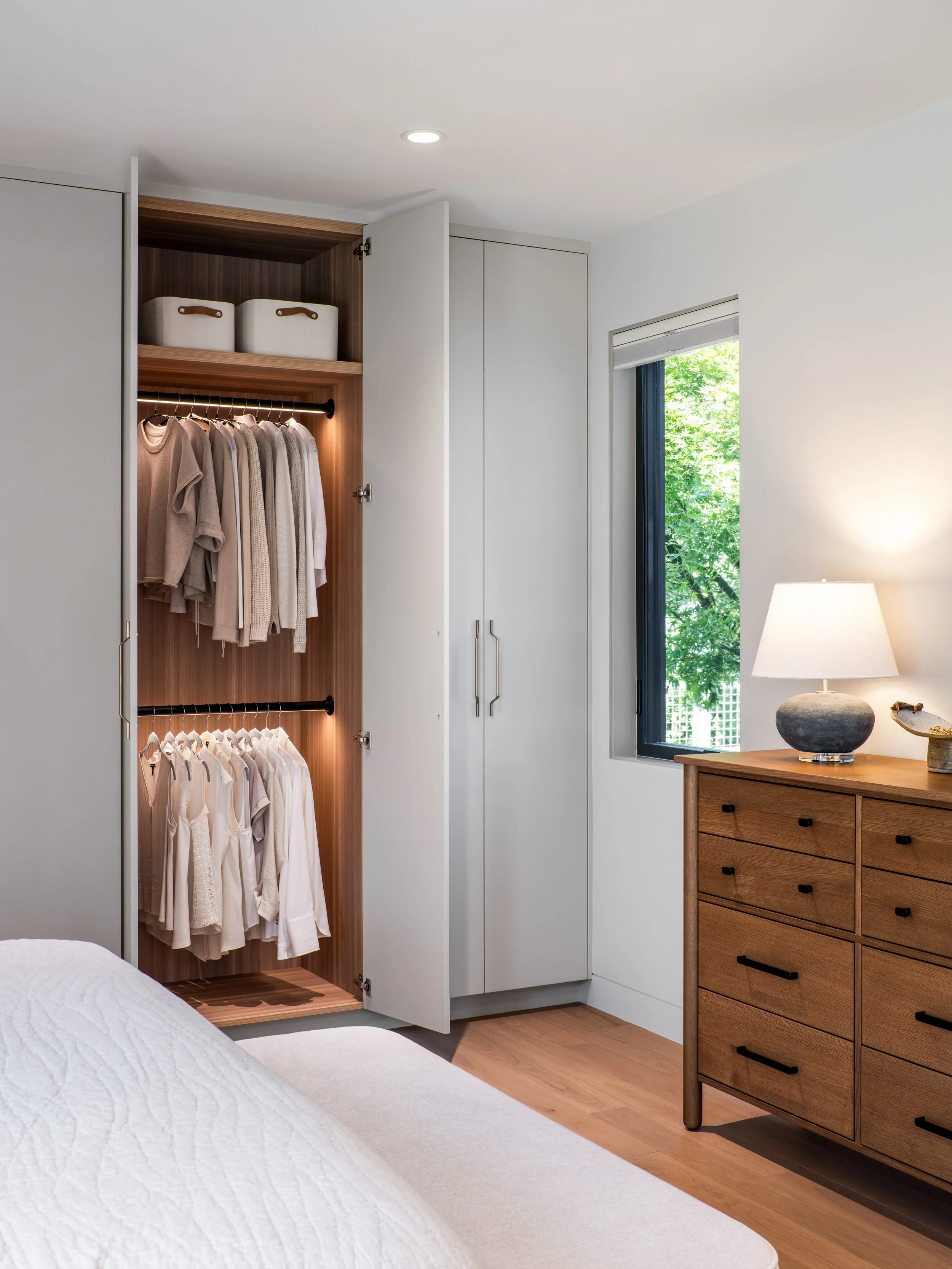 A neatly organized closet with a few beige and white clothes hanging on a black rod, two white storage baskets on the top shelf, and a window with a view of greenery outside. There is a wooden dresser with a lamp on it in a well-lit bedroom.