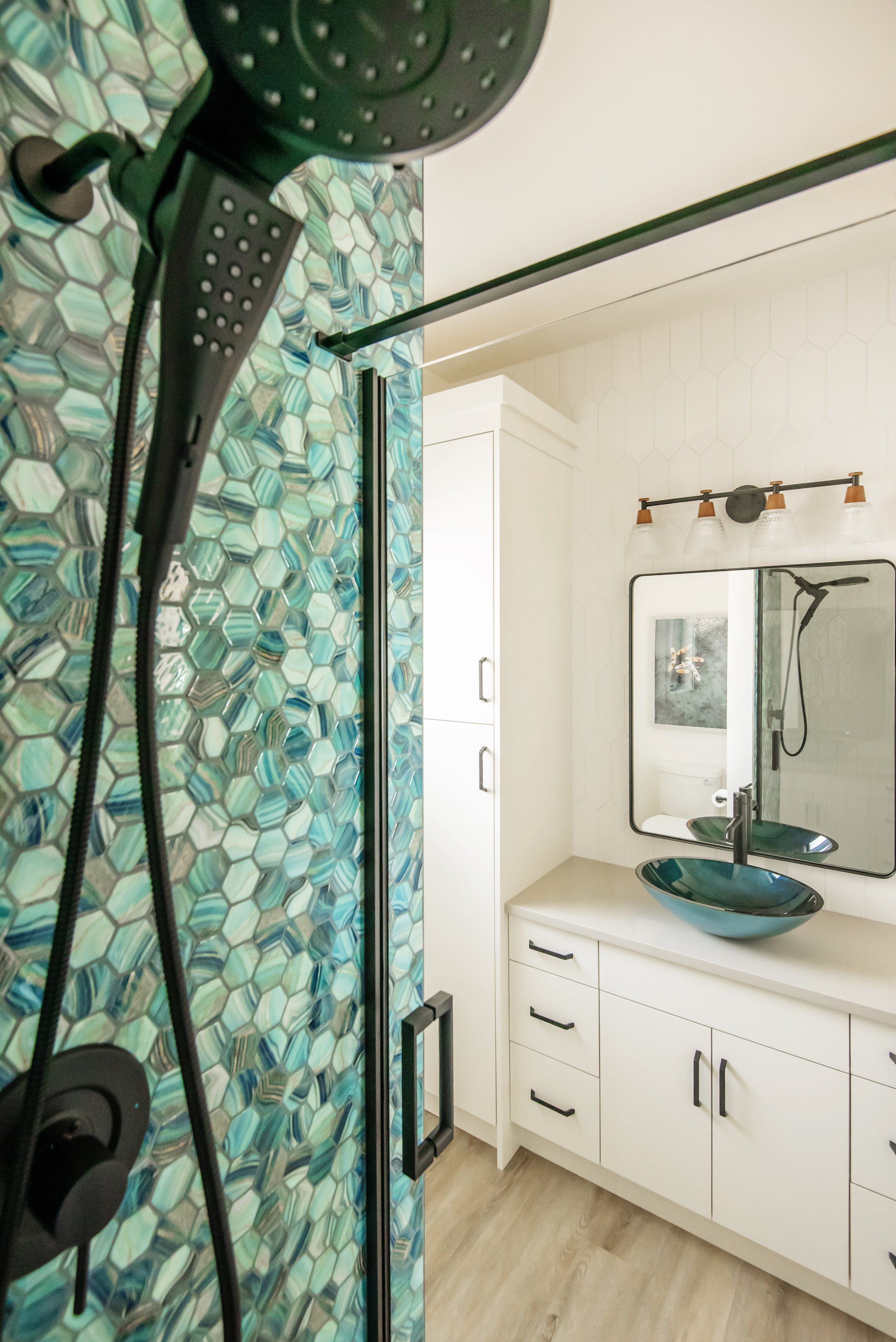 A bathroom with a mirror, navy sinks, white cabinets, and a shower with a colorful tiled wall.