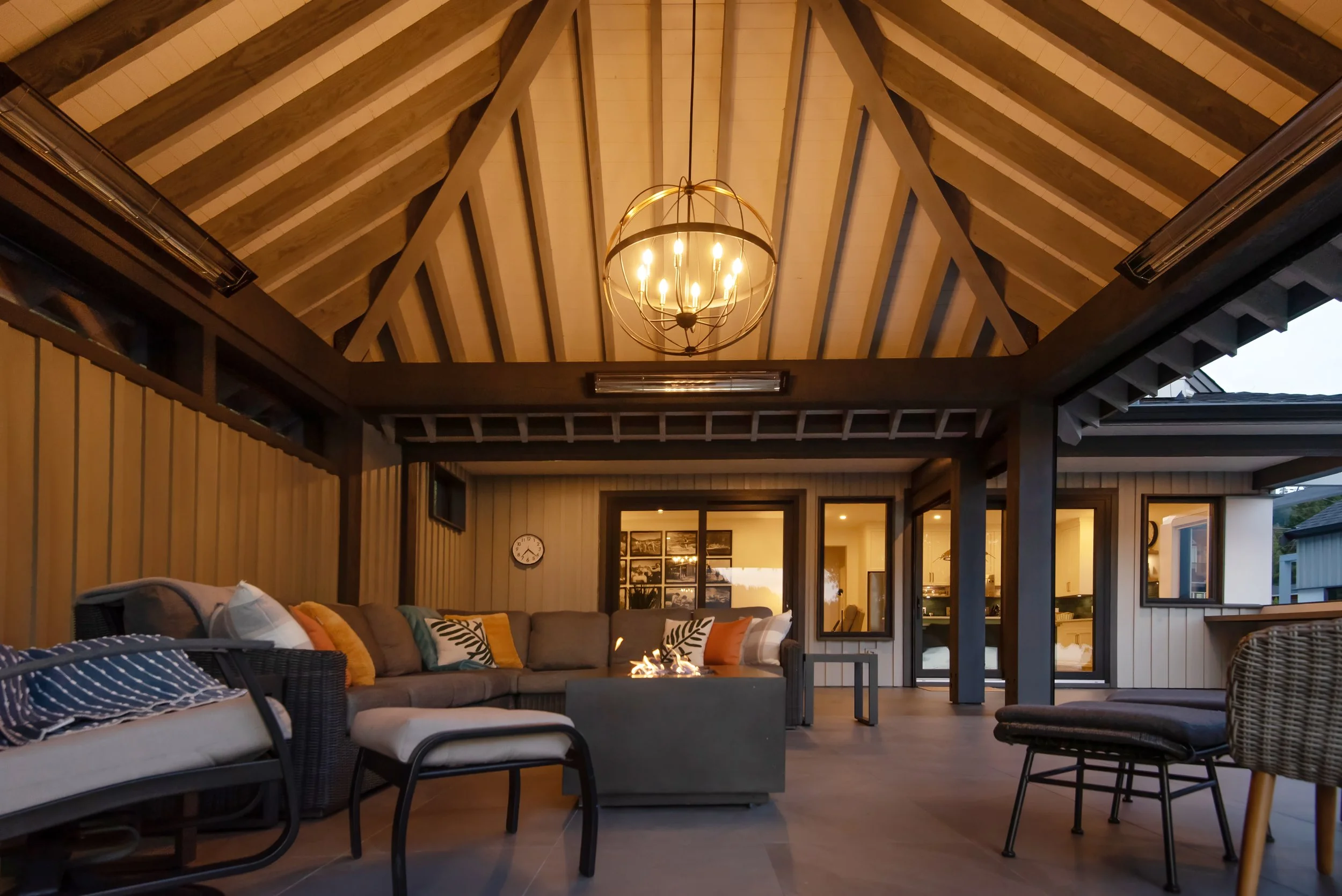 Covered outdoor living space with a chandelier hanging from a vaulted wood ceiling. There's a firepit, outdoor furniture including a sectional sofa with colorful cushions, a wicker chair, and a lounge chair. The space is illuminated with warm lighting, and there's a wall clock and framed pictures on the wall.