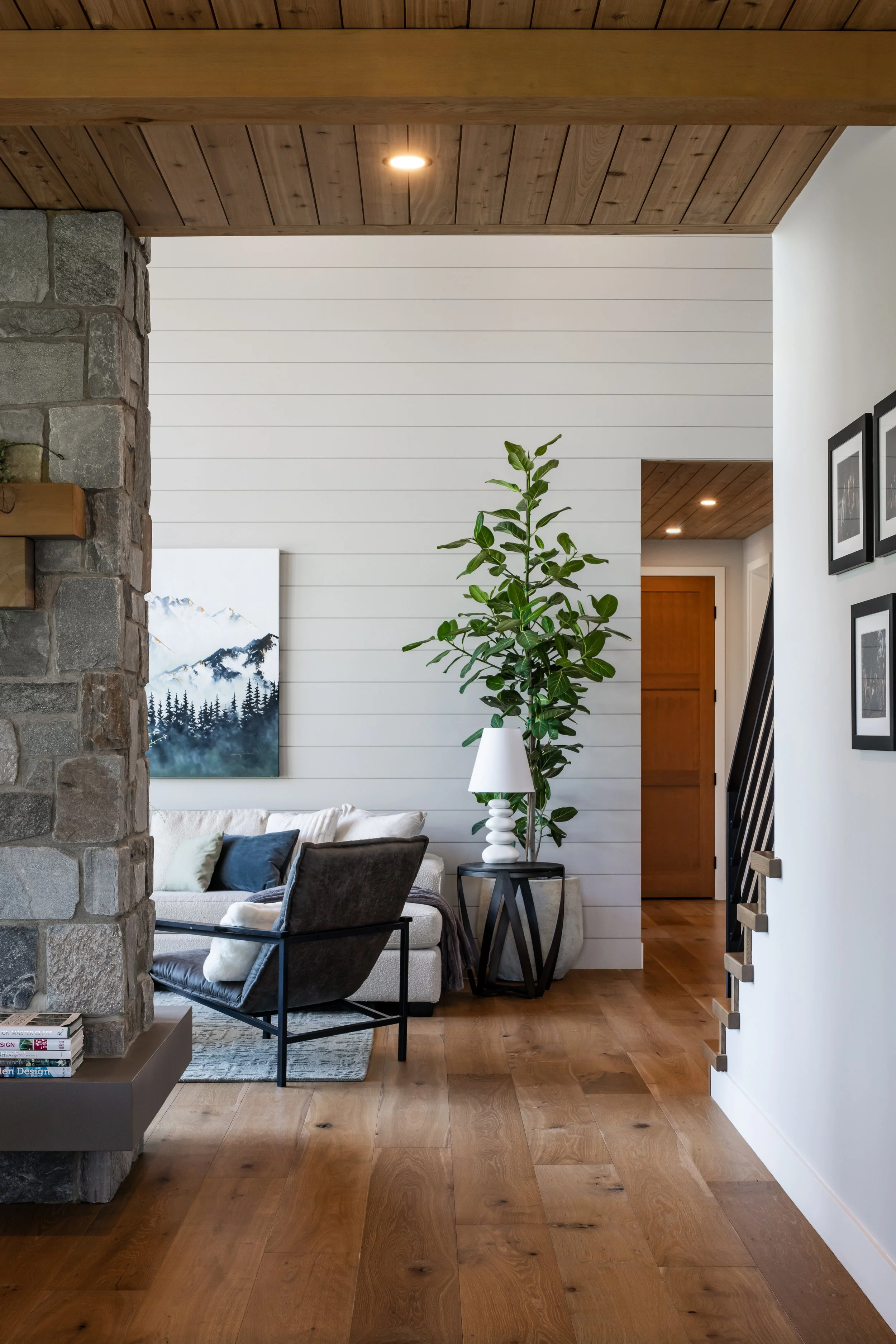 Living room area with a stone wall on the left, a white shiplap wall in the background, and a wooden ceiling. There are various chairs and a sofa with pillows, a side table with a white lamp, a large green plant in a white pot, and framed pictures on the wall to the right. A mountain landscape painting is partially visible on the left.