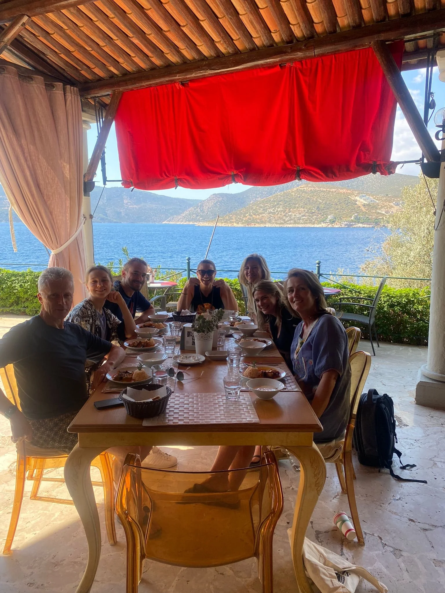 A group of eight people sitting around a dining table with food, near a lake or ocean, with mountains in the background and a red curtain overhead.