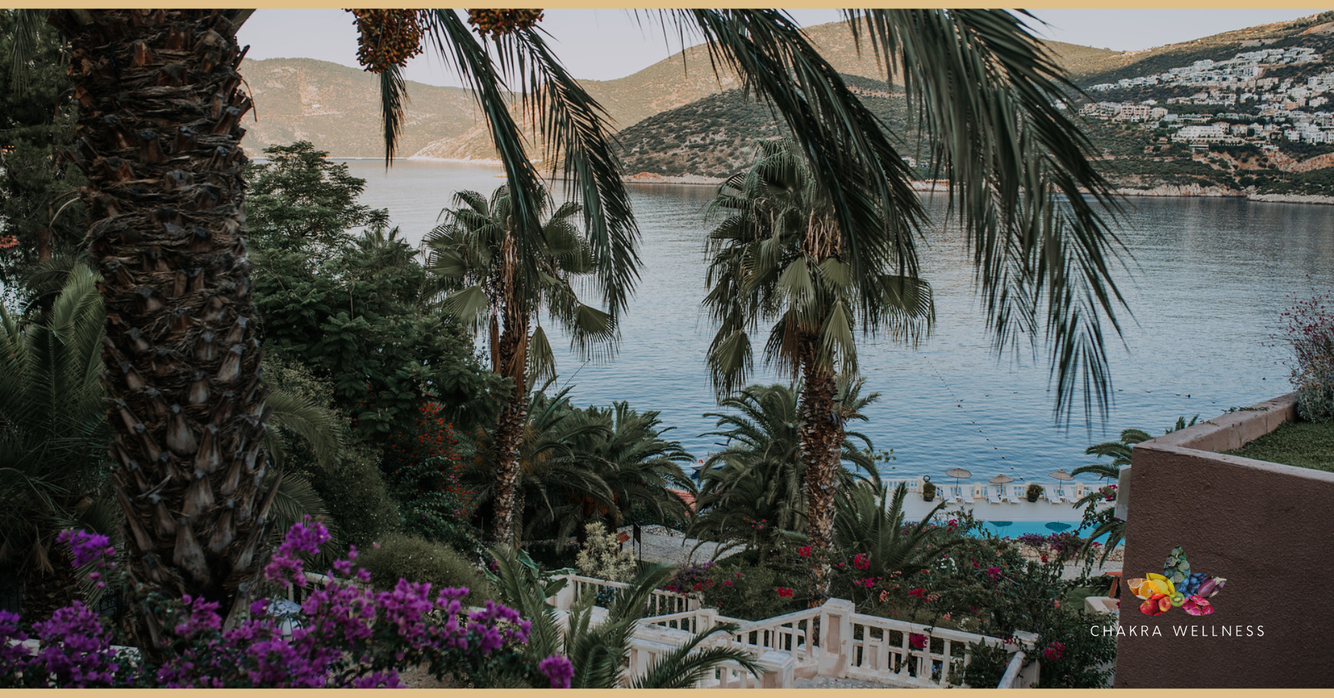 Scenic view of tropical landscape with palm trees, colorful flowers, a swimming pool, and a body of water in the background, with hilly terrain.