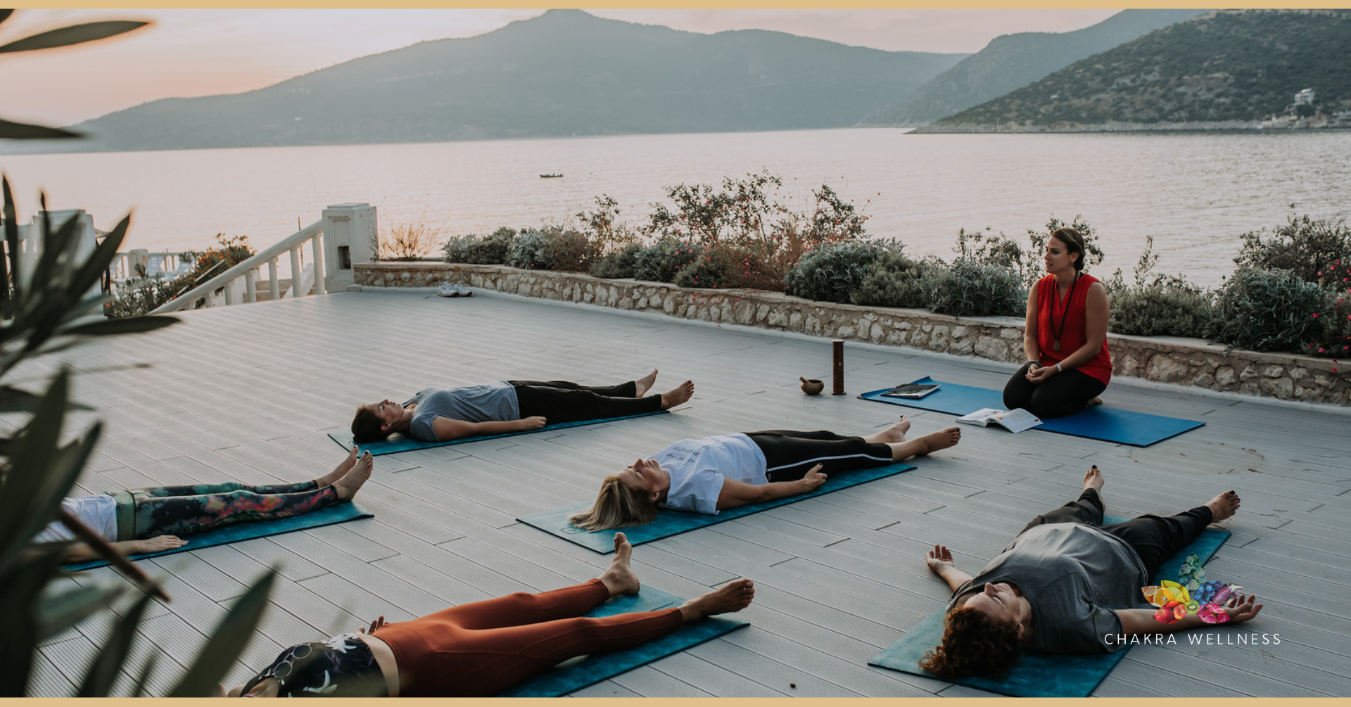 Group of people practicing yoga outdoors on a deck by a lake with mountains in the background, led by an instructor sitting cross-legged in front.