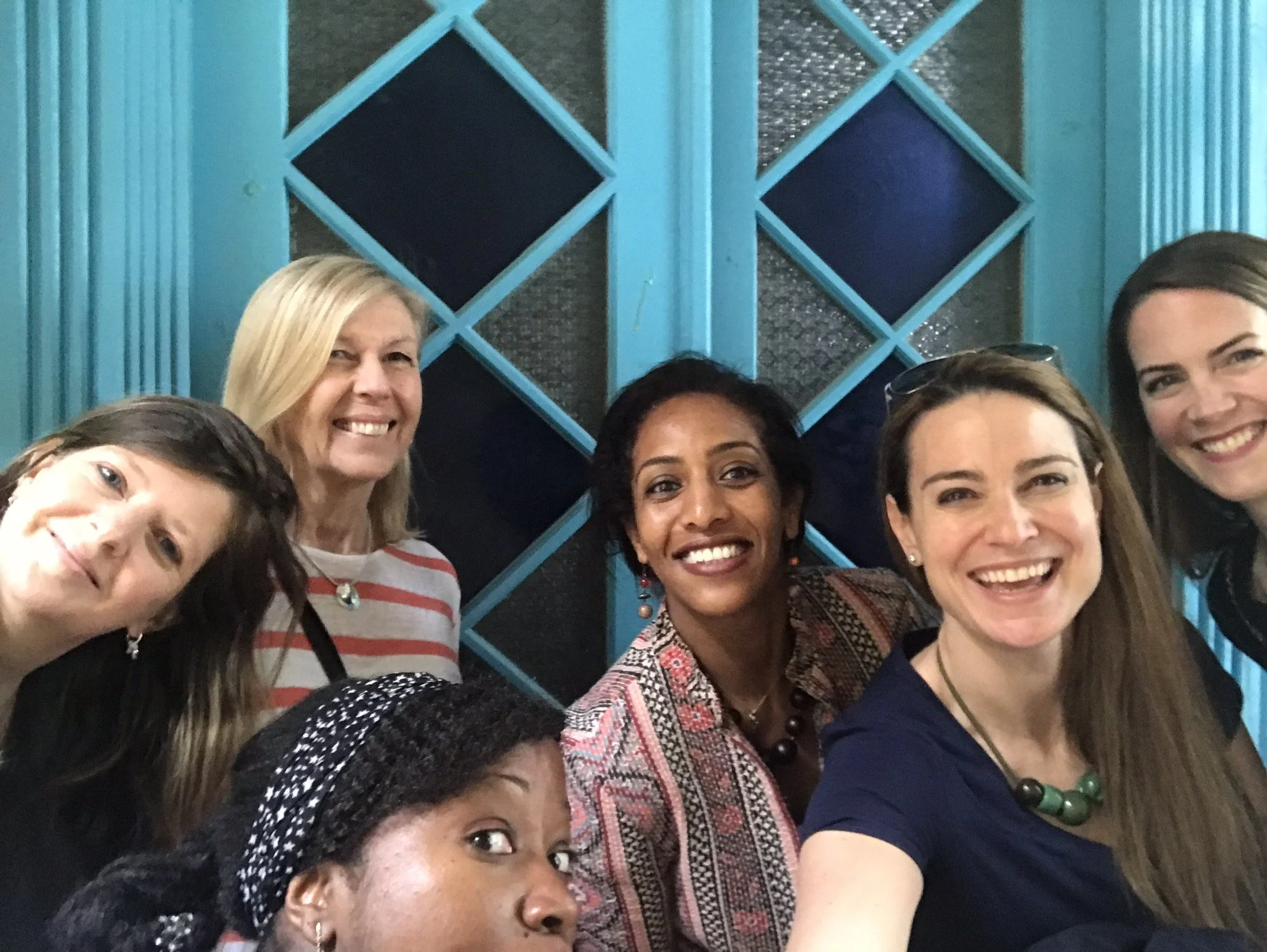 Six women smiling for a group photo indoors in front of a decorative blue wall with diamond-shaped and rectangular panels.