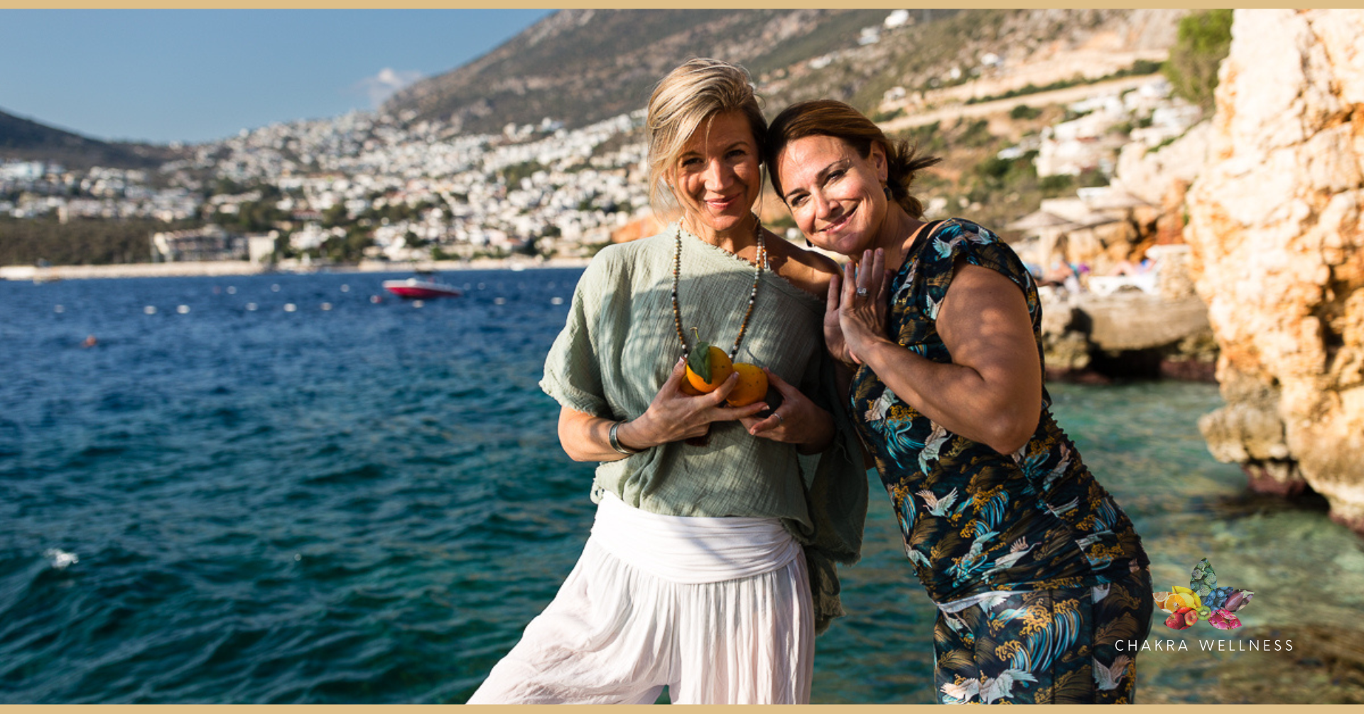Two women smiling and posing by the water with a scenic coastal background, holding a small fruit or vegetable.