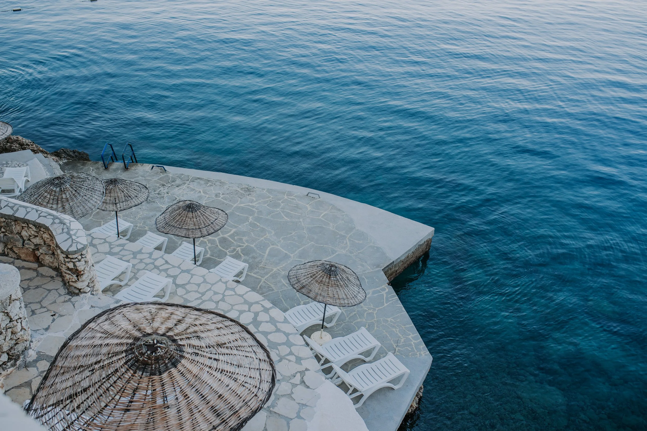 Empty poolside area with white lounge chairs, bamboo umbrellas, and a ladder leading into the water, overlooking the ocean.